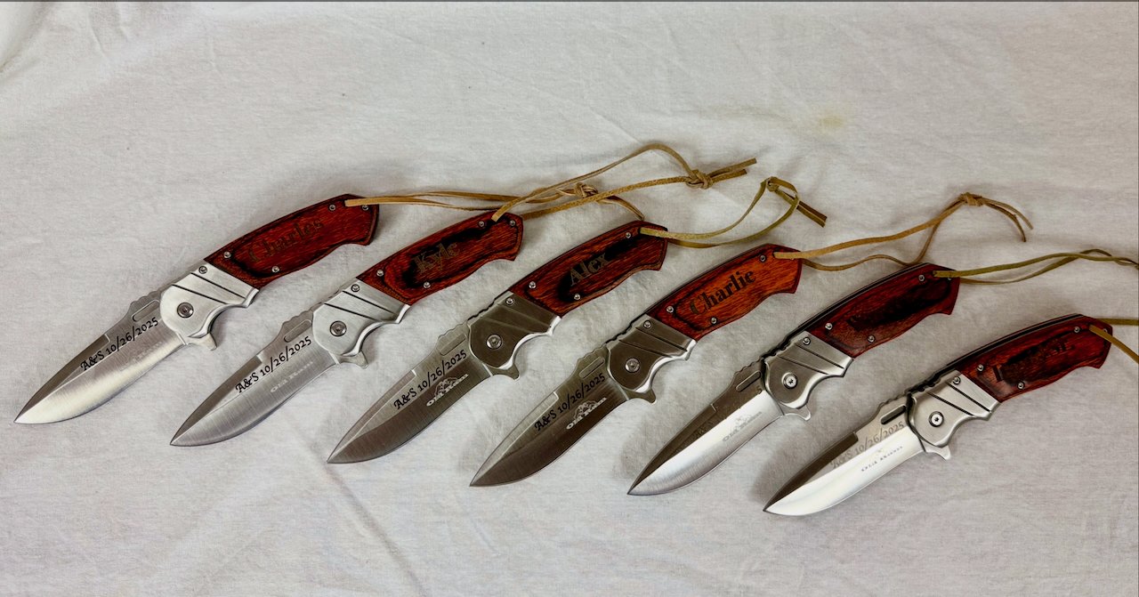 Six folding knives with wooden handles, arranged in a row on a white surface, some inscribed with names and a date.