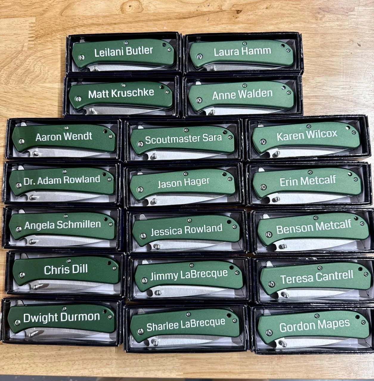 A collection of 15 name tags with green background, arranged in five rows on a wooden surface. Each name tag bears a different person's name.