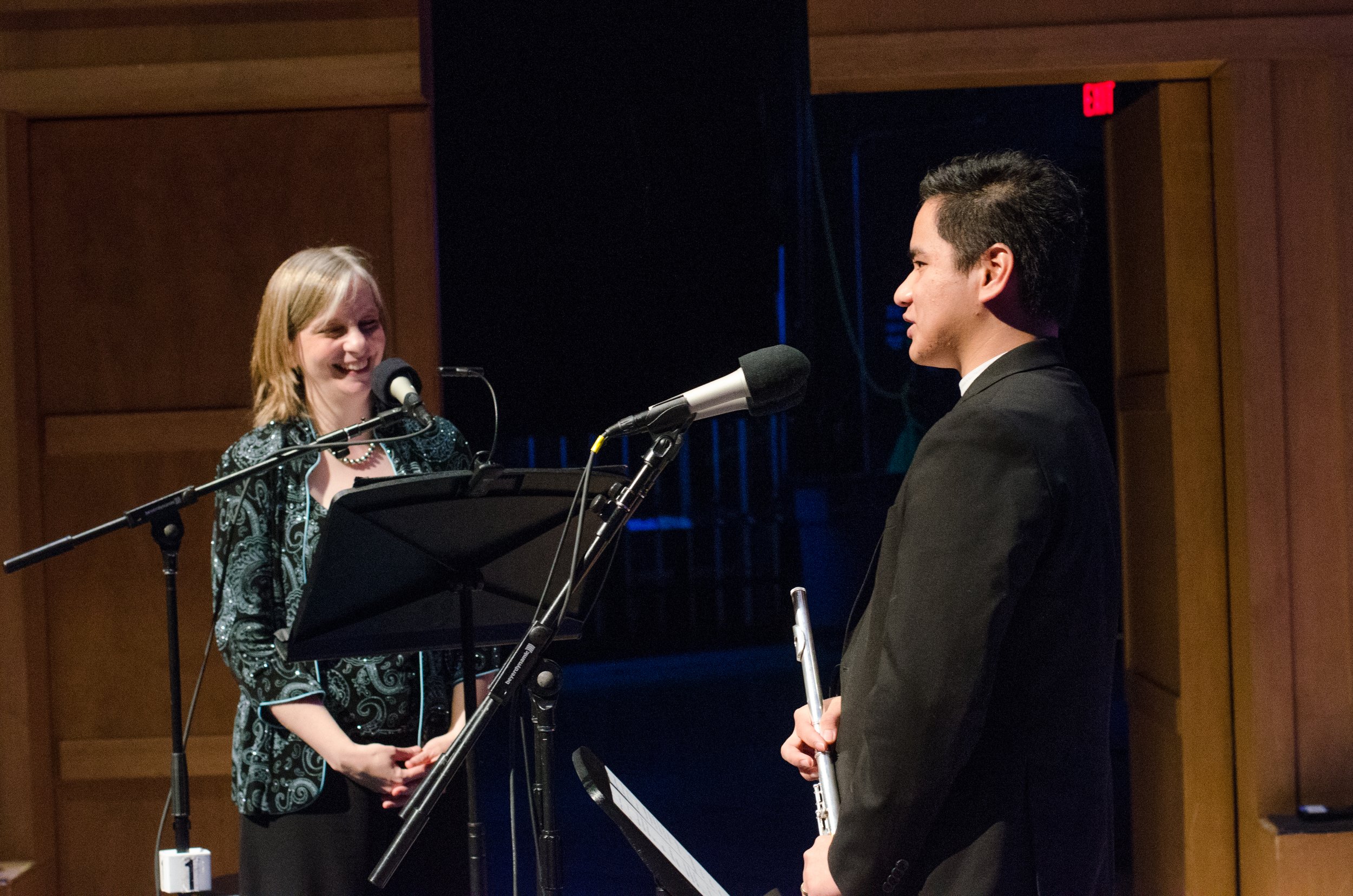 A woman and a man standing in front of microphones on a stage, with the woman smiling and the man holding a flute.