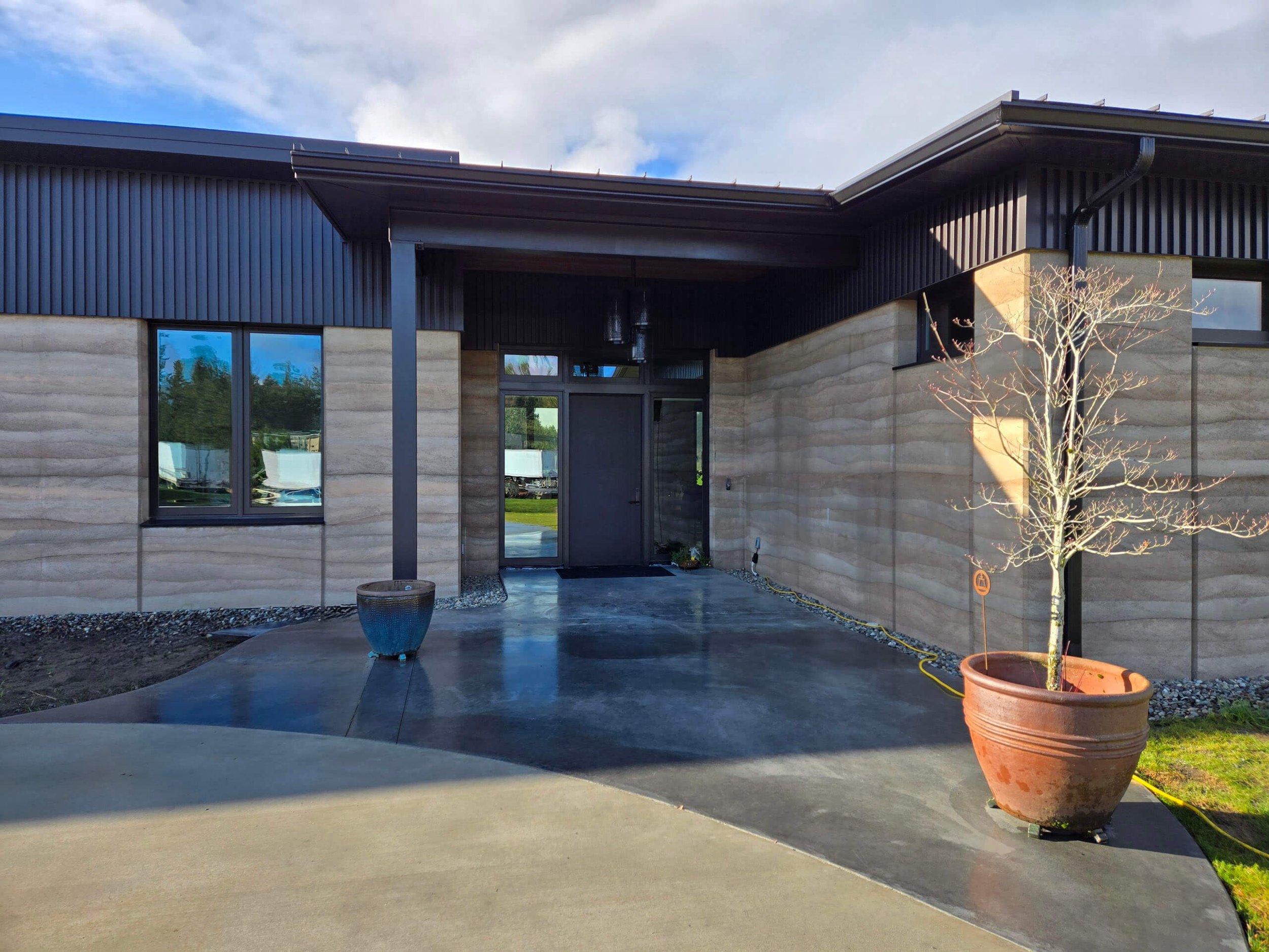 Modern house with black metal siding and a concrete walkway, potted trees, and a reflective glass door.