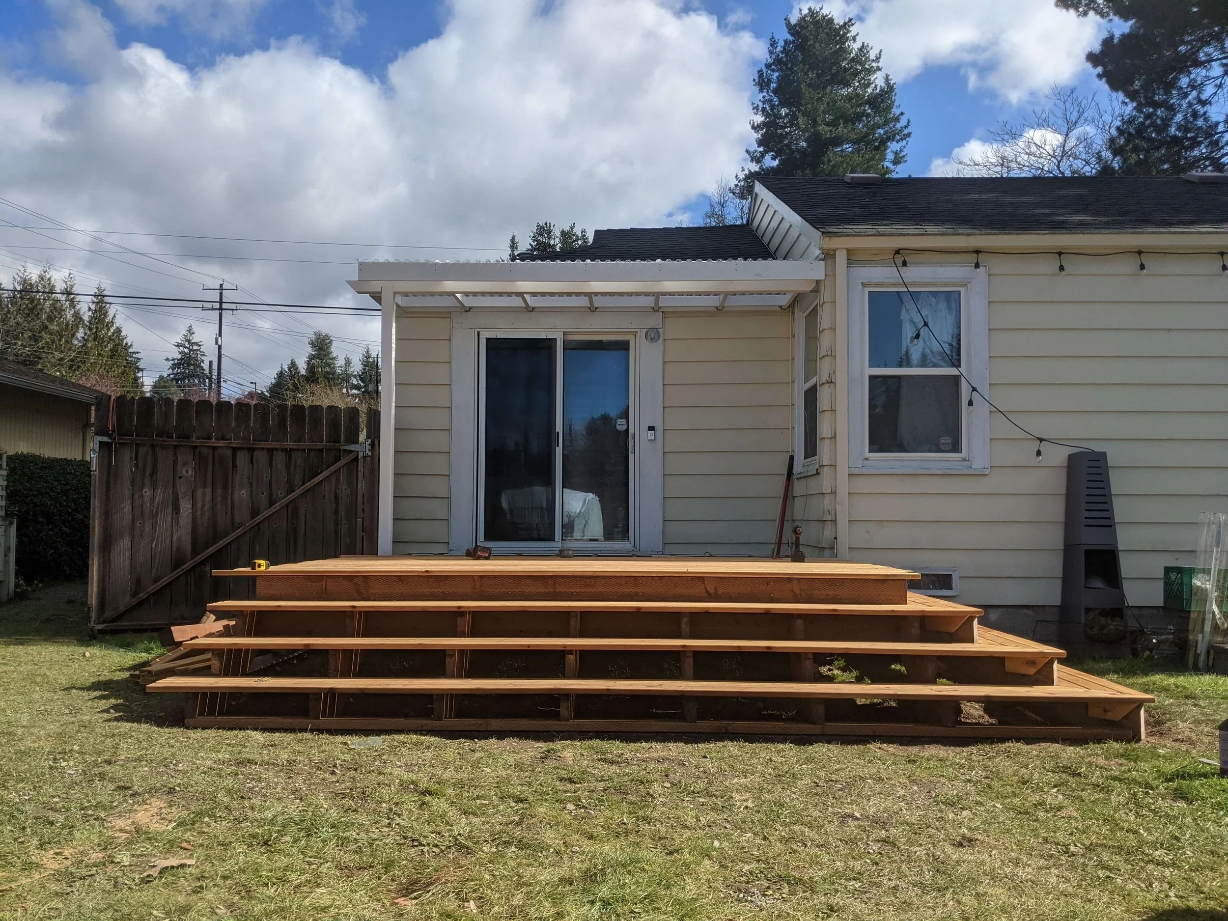 Back of a house with a yellow exterior, a sliding glass door, and a window, with a wooden deck under construction with stairs leading up to the deck.