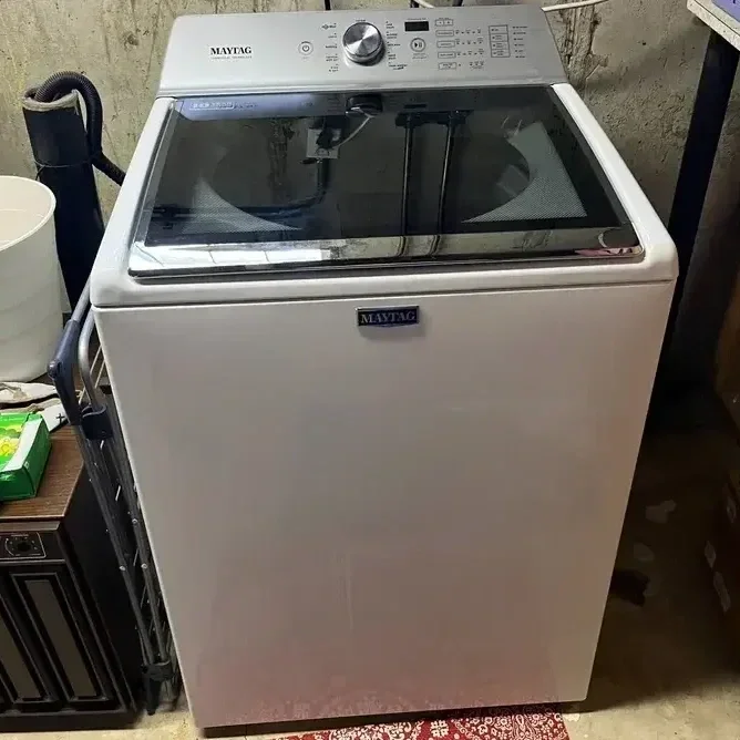 White top-loading washing machine with a black control panel and lid, positioned in a utility area.