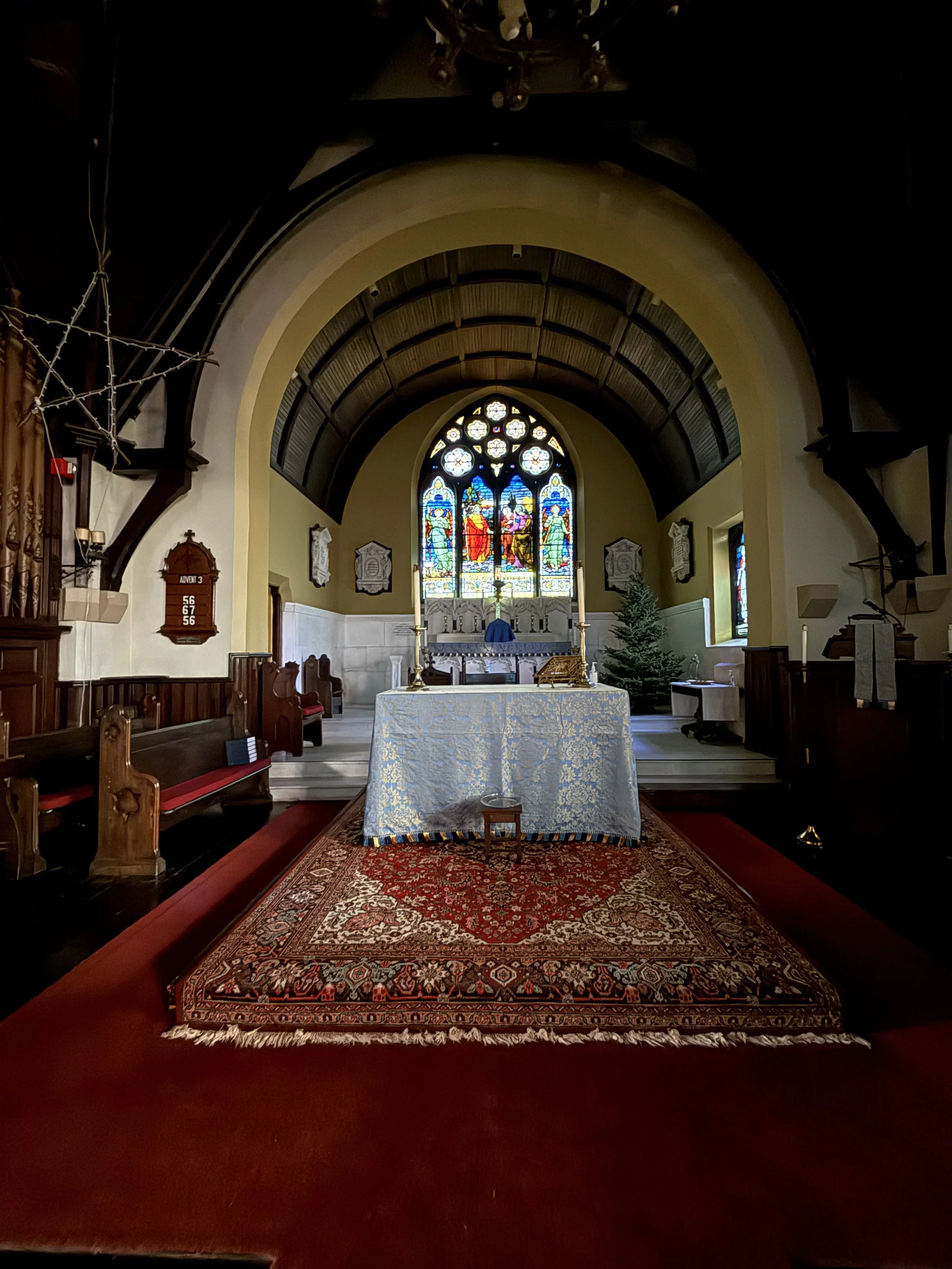 Interior of a church showing the altar with a blue cloth, decorative rug, stained glass windows in the background, pews on either side, and a high arched ceiling.