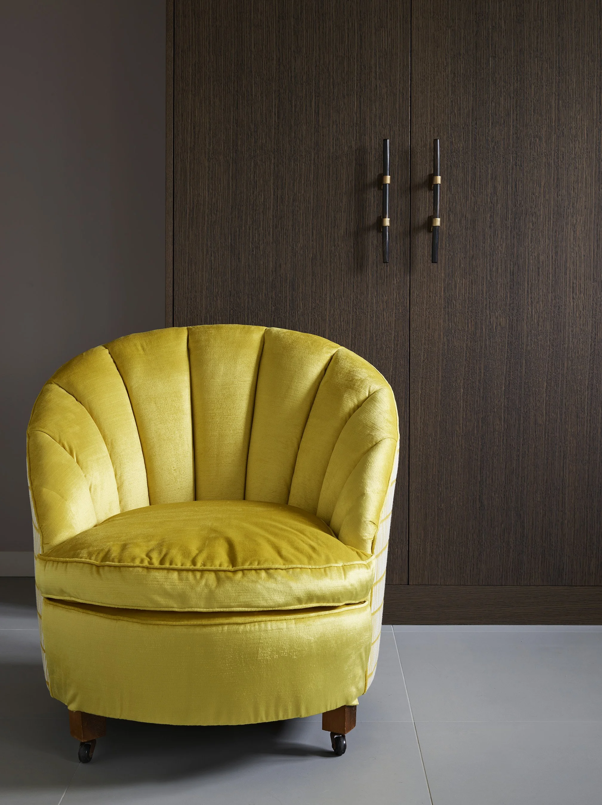 A yellow upholstered armchair with wooden legs and caster wheels, placed on a light-colored tiled floor, against a dark wood-paneled wall with black and gold vertical handles.