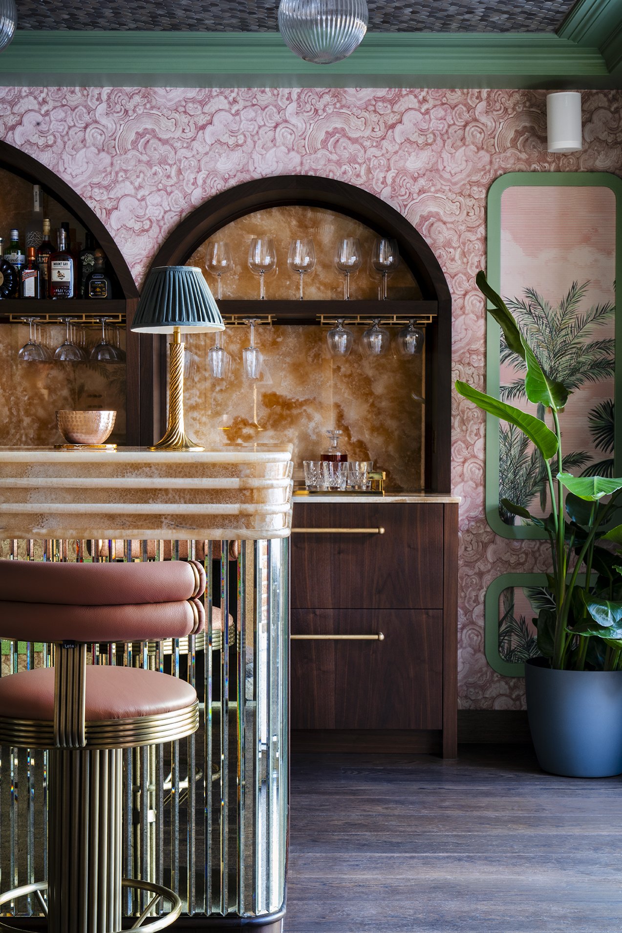 Pink and green interior of a bar with a marble counter, a pink bar stool with gold accents, a lamp with a teal shade, wooden shelves with wine glasses, pink patterned walls, a tall potted green plant, and a dark wood floor.
