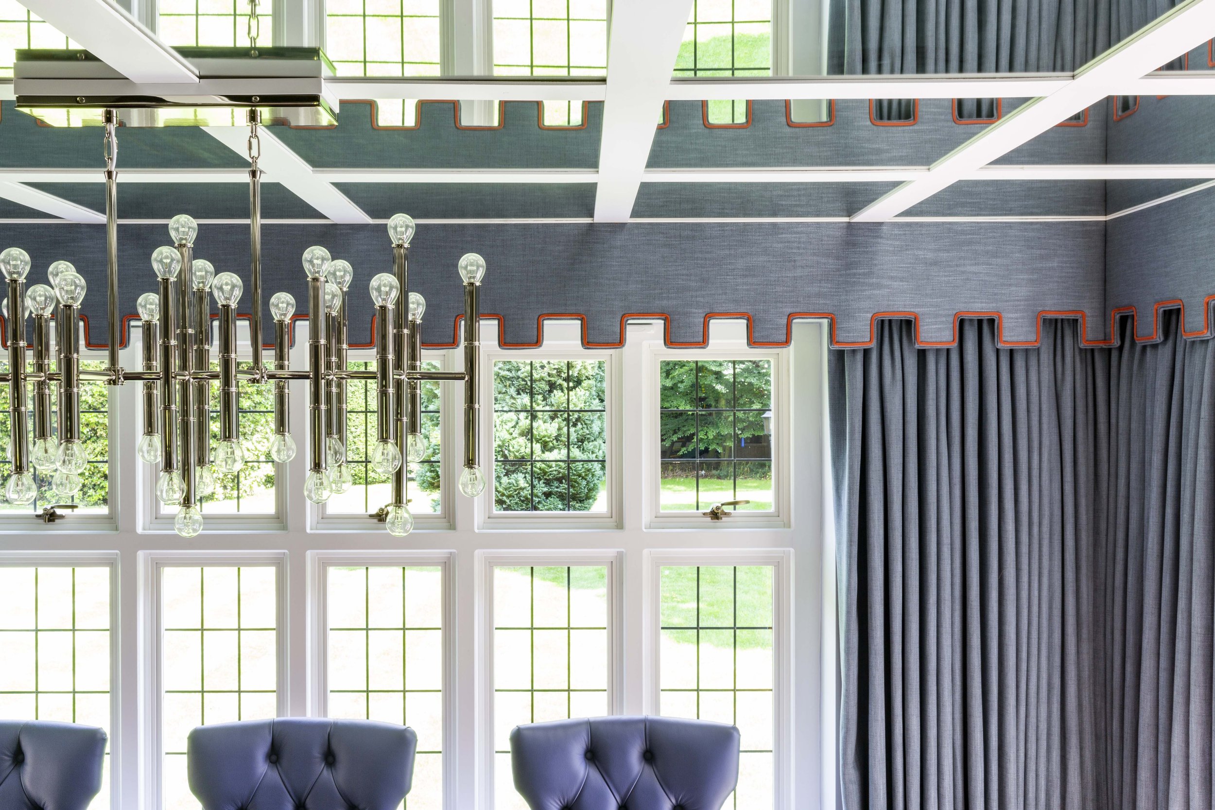 Interior room with large windows, blue curtains with red trim, modern chandelier with hanging light bulbs, and gray upholstered chairs.