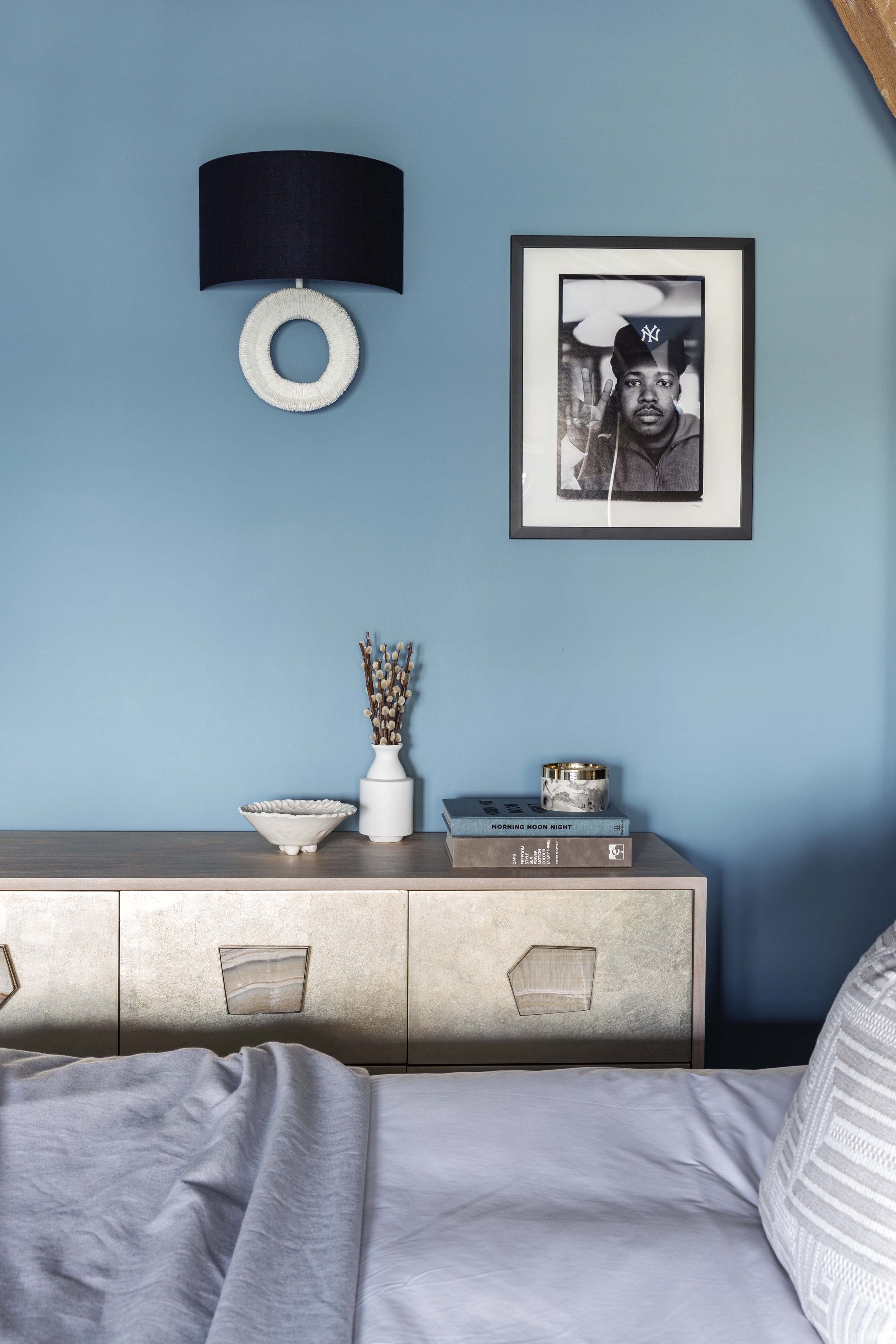 A bedroom with a blue wall, a framed black-and-white photo of a man wearing a New York Yankees hat, a black lampshade with a white circular wall decoration, a wooden dresser with decorative items, and a bed with white linens and a striped pillow.