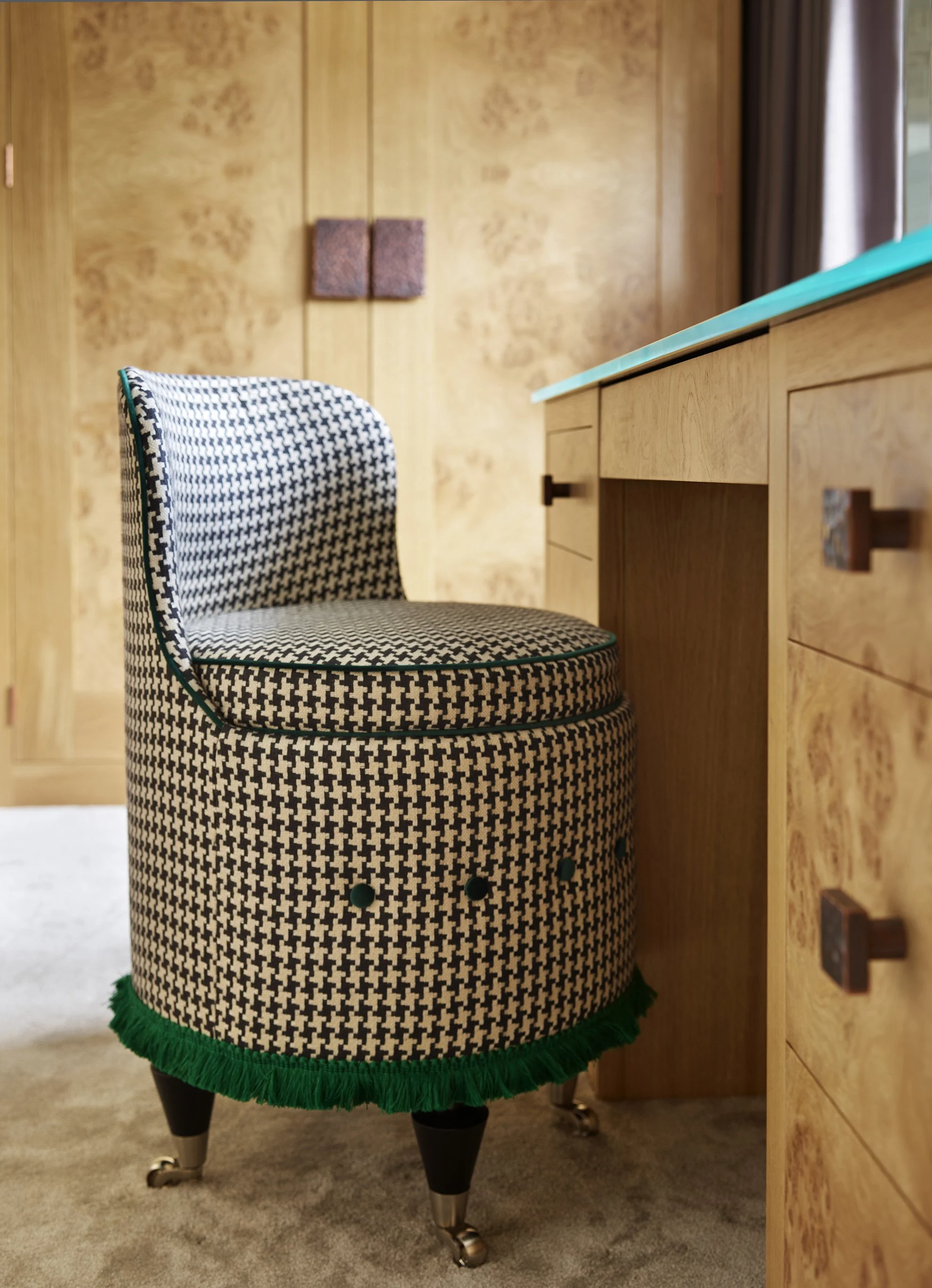 A vintage-style chair with black and white houndstooth pattern, green trim, and wooden legs with casters, placed beside a wooden desk in a room with wood-paneled walls.