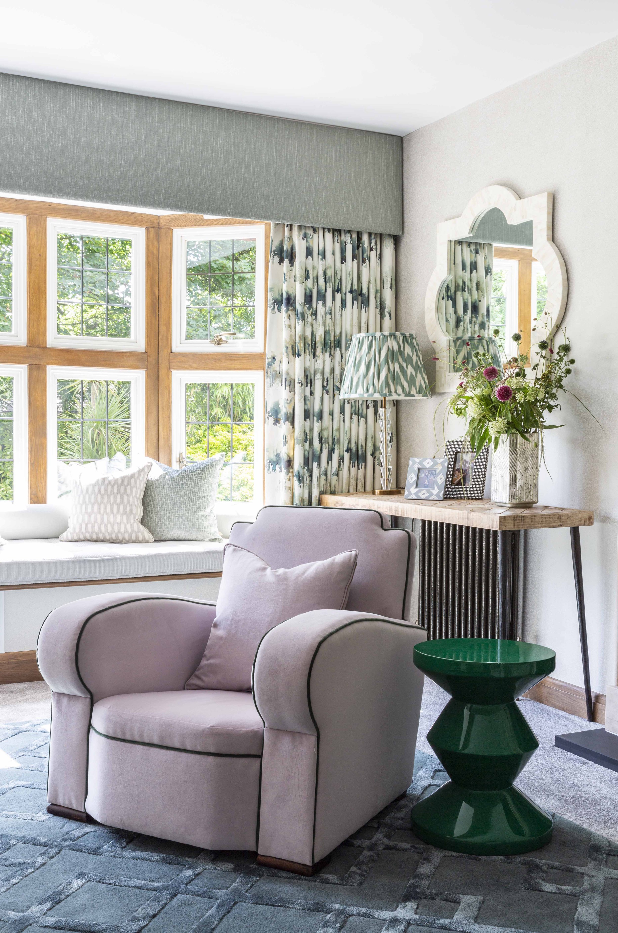 A cozy living room corner with a large window seat with pillows, a cream armchair with a pillow, a wooden side table with framed photos, a table lamp with a patterned shade, a vase with flowers, a green side table, a mirror on the wall, and curtains with a floral pattern.