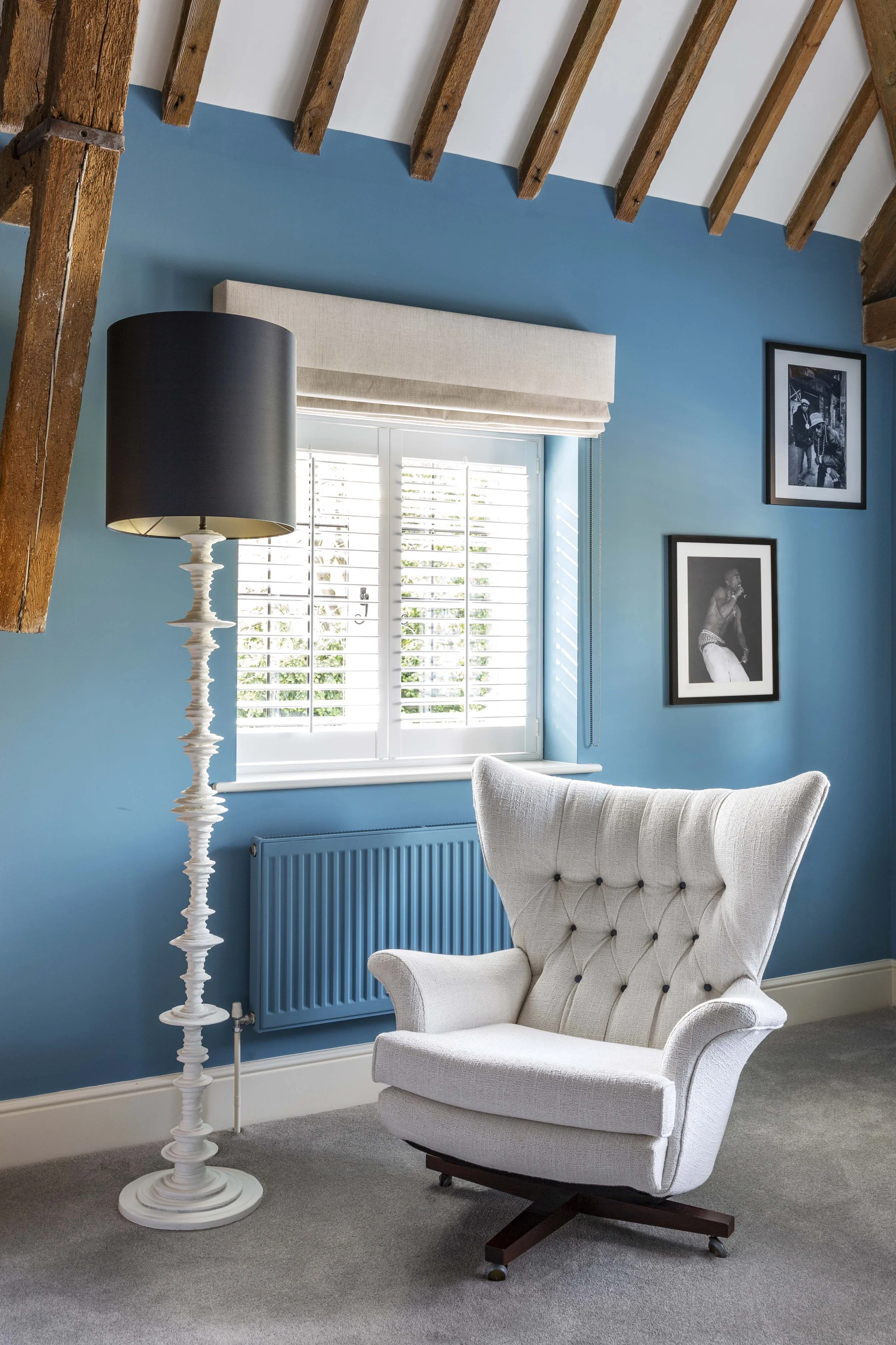 A cozy room with a blue accent wall, a white armchair, a tall white floor lamp with a black shade, a window with white shutters, framed black-and-white photos on the wall, and an exposed wooden beam ceiling.