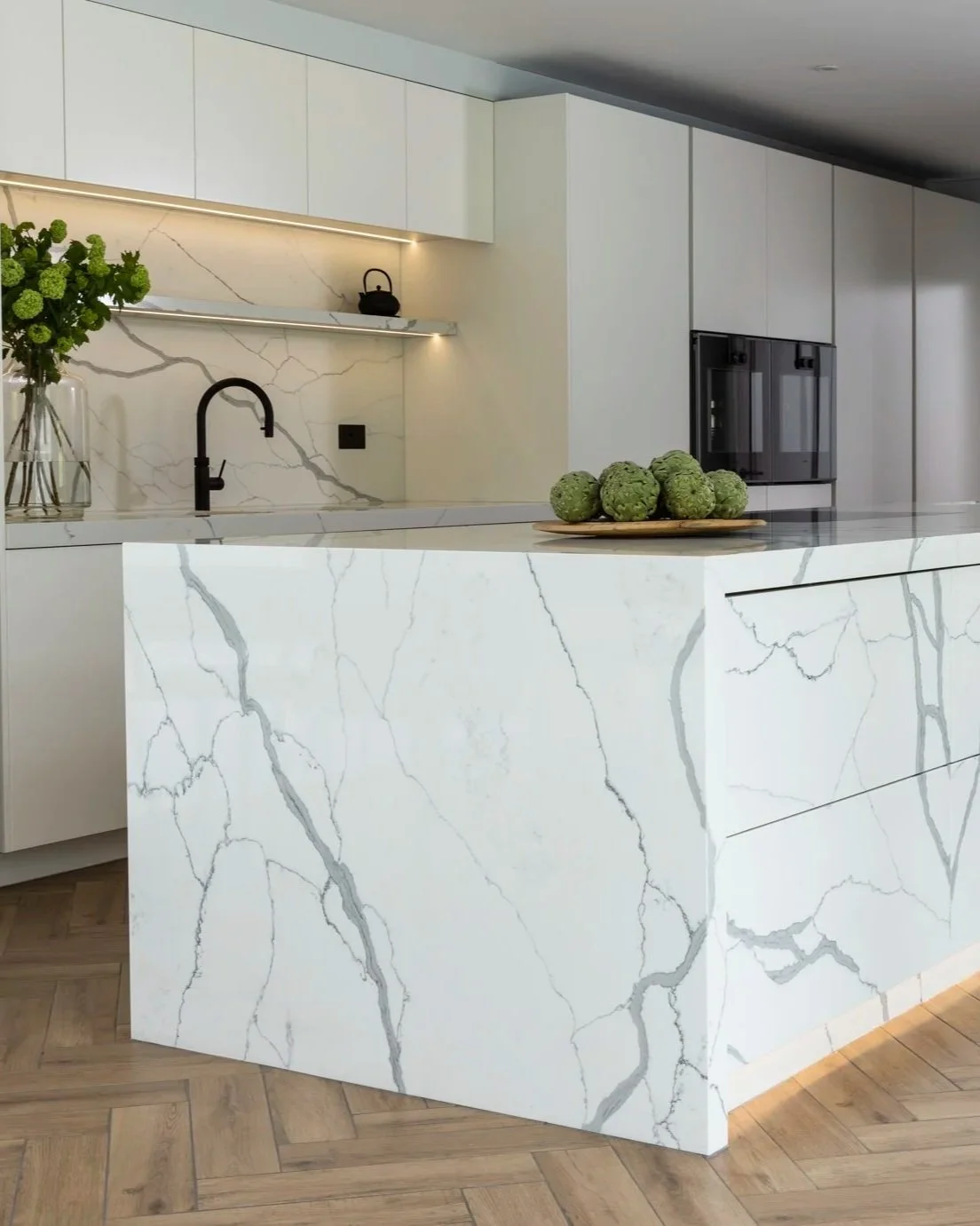 Modern kitchen with a white marble island featuring gray veins, a black faucet, white cabinets, and a wooden tray with green artichokes on the counter.