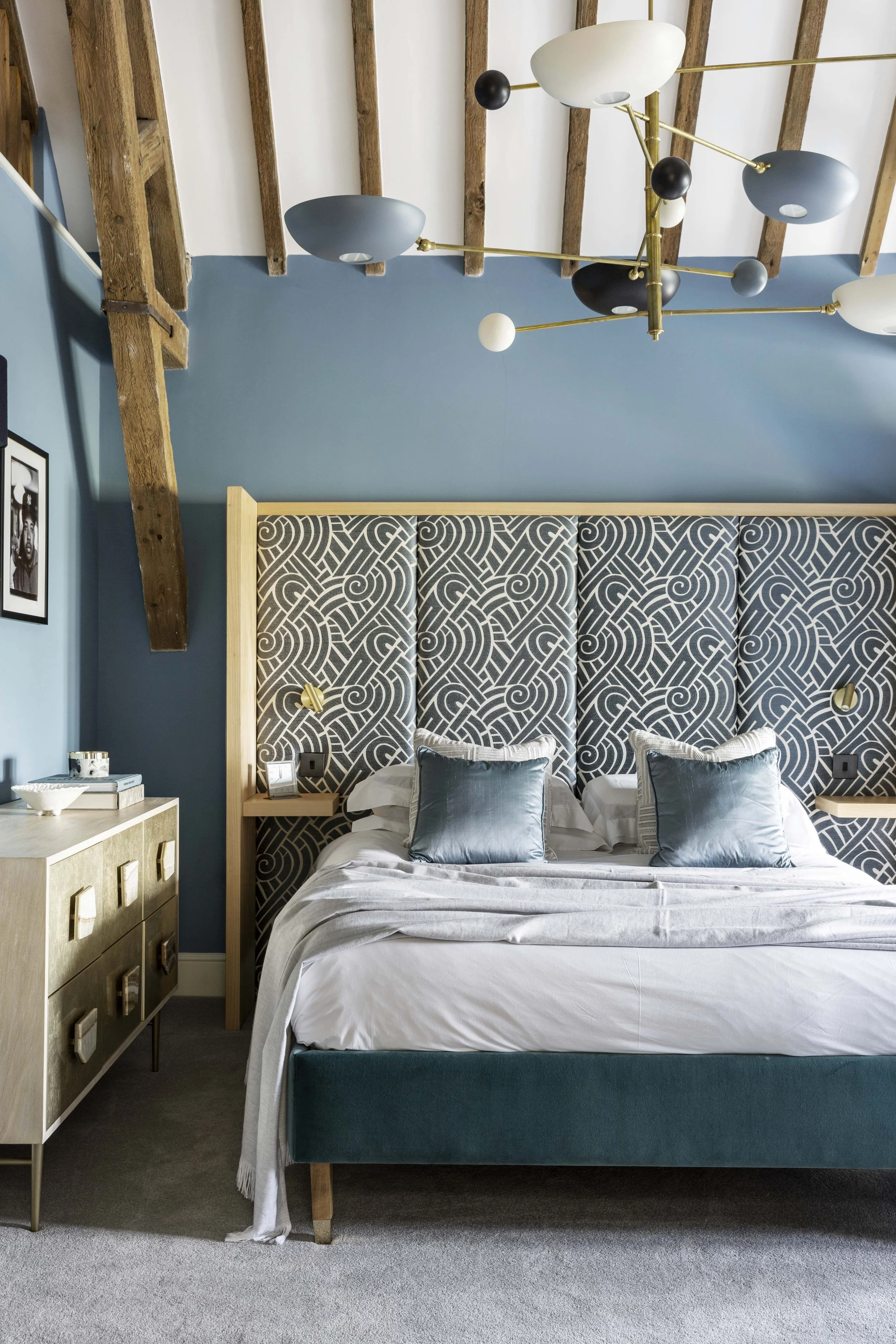 A modern bedroom featuring a bed with a patterned headboard, blue pillows, a dresser, a framed photo on the wall, and a multi-colored chandelier hanging from a wooden ceiling.