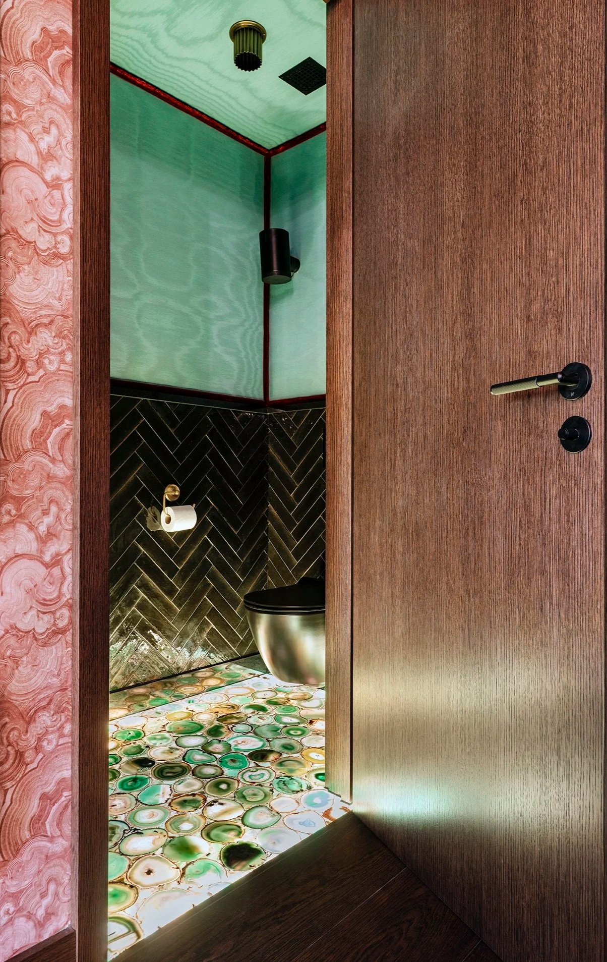 Interior view of a stylish bathroom with black chevron tile walls, a gold toilet, a paper holder, and an illuminated floor with colorful agate stone patterns.