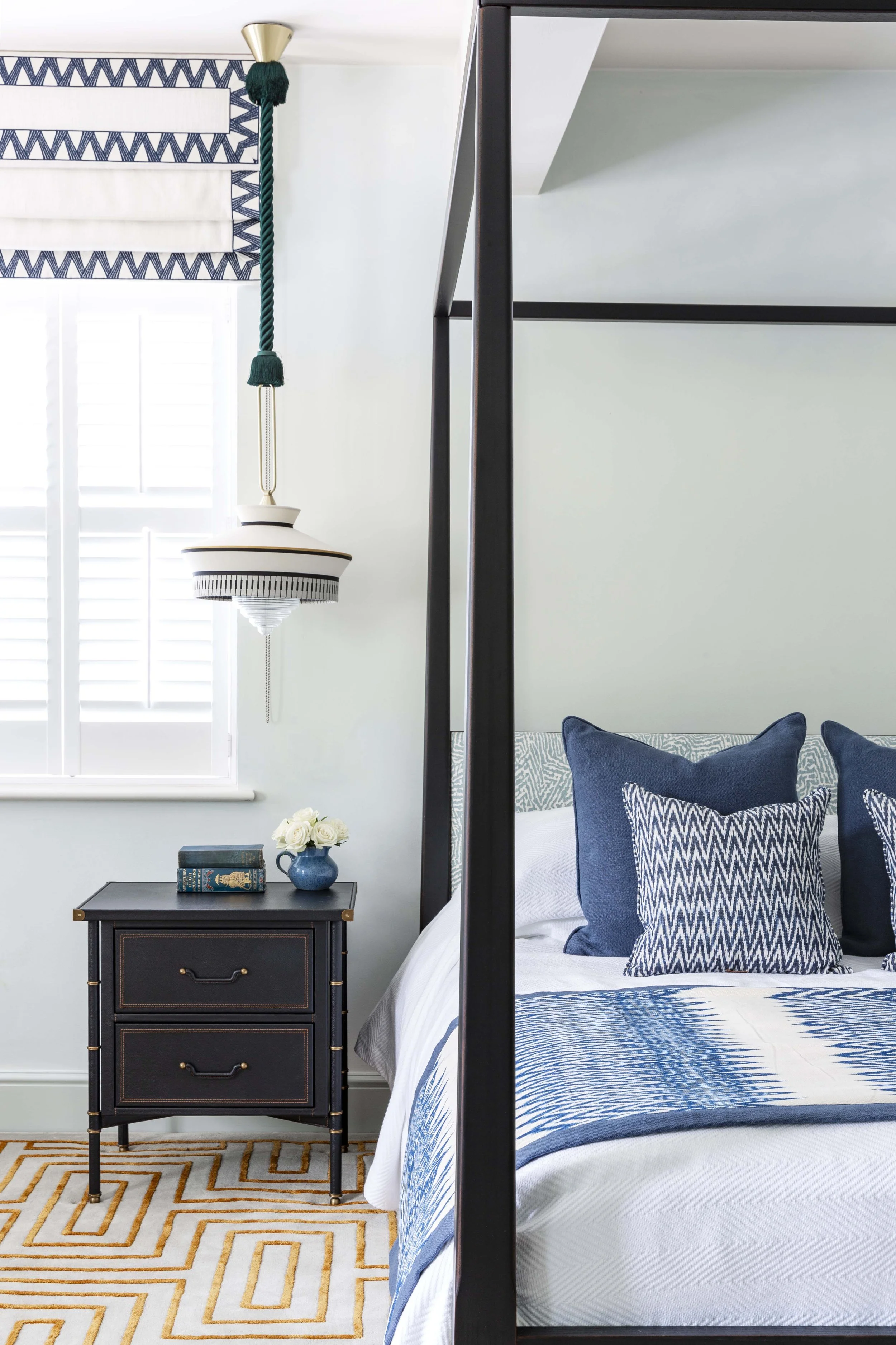 A bedroom with a black metal four-poster bed, blue and white pillows, and a quilt. Next to the bed, there is a black nightstand with two drawers, a blue vase with white roses, and two stacked books. There is a window with white shutters and a patterned blue and white roman shade. A ceiling light with a decorative pull chain hangs from the ceiling.