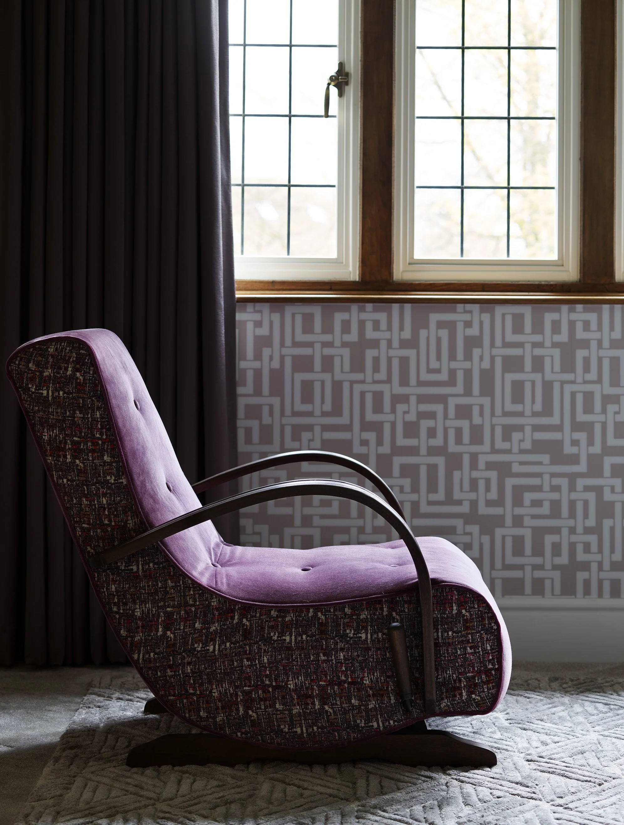 A purple upholstered armchair with a patterned backrest and curved armrests in front of a window with grid panes, dark curtains, and a geometric-patterned wall behind.