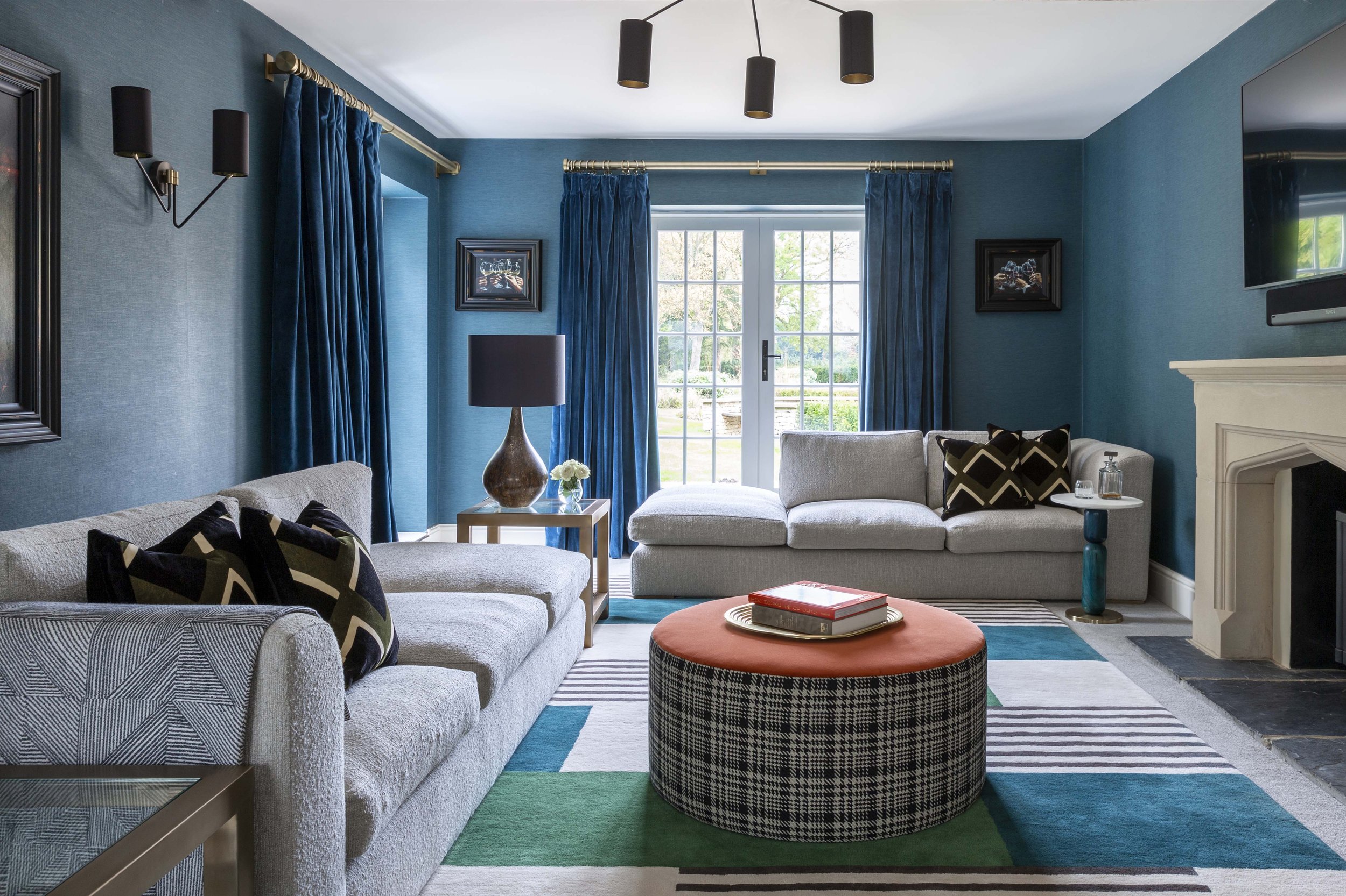A contemporary living room with blue walls, large windows with blue curtains, two light-colored sofas with patterned pillows, a round ottoman with a red top and black-and-white checkered base, a black table lamp on a side table, and a fireplace with a wall-mounted TV above.