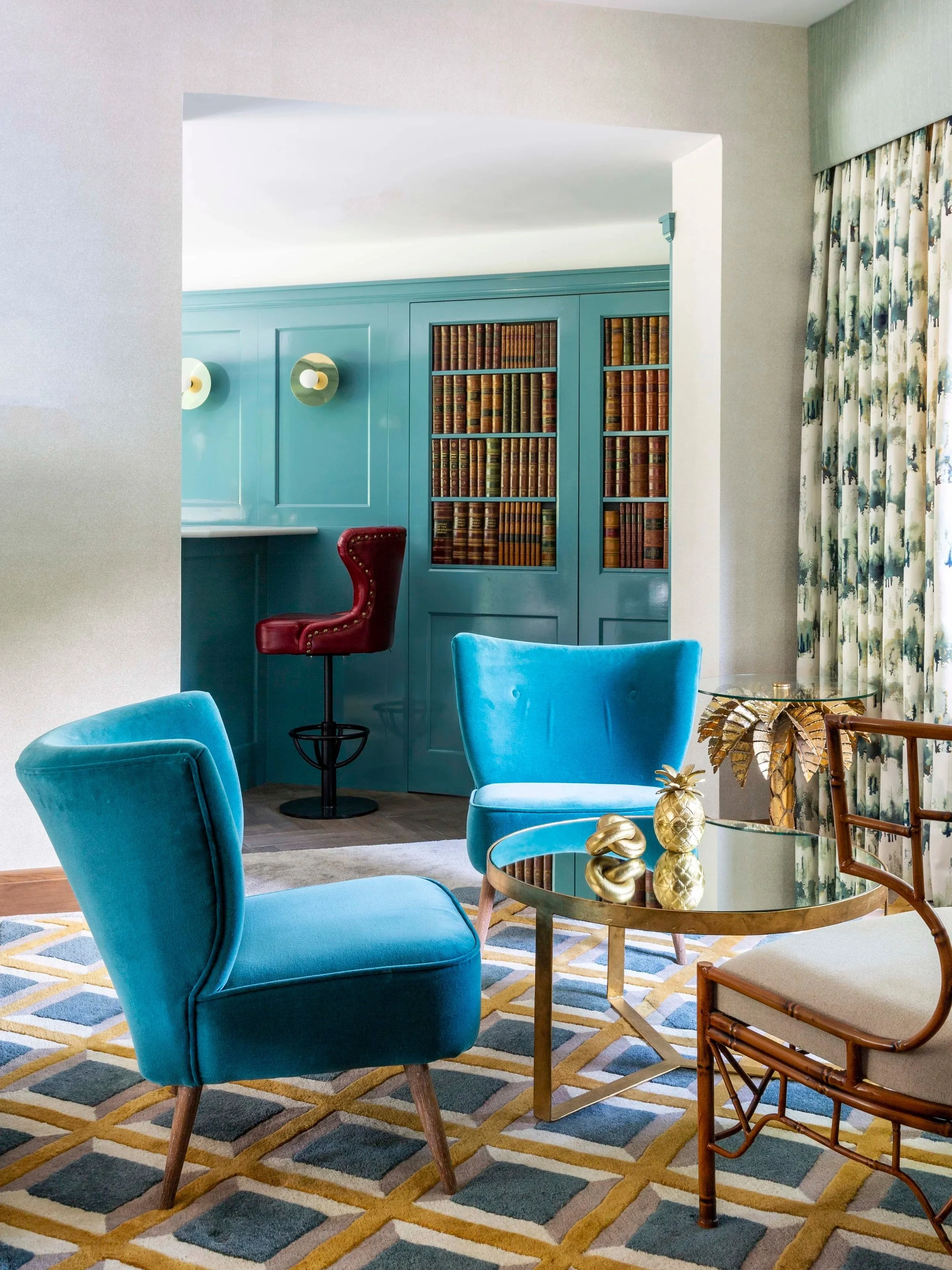 Living room with blue velvet chairs, a geometric patterned rug, and a gold coffee table with decorative gold pineapple and sculptures. In the background, there is a built-in bookshelf with leather-bound books, a teal bar area with a red leather barstool, and floral curtains.