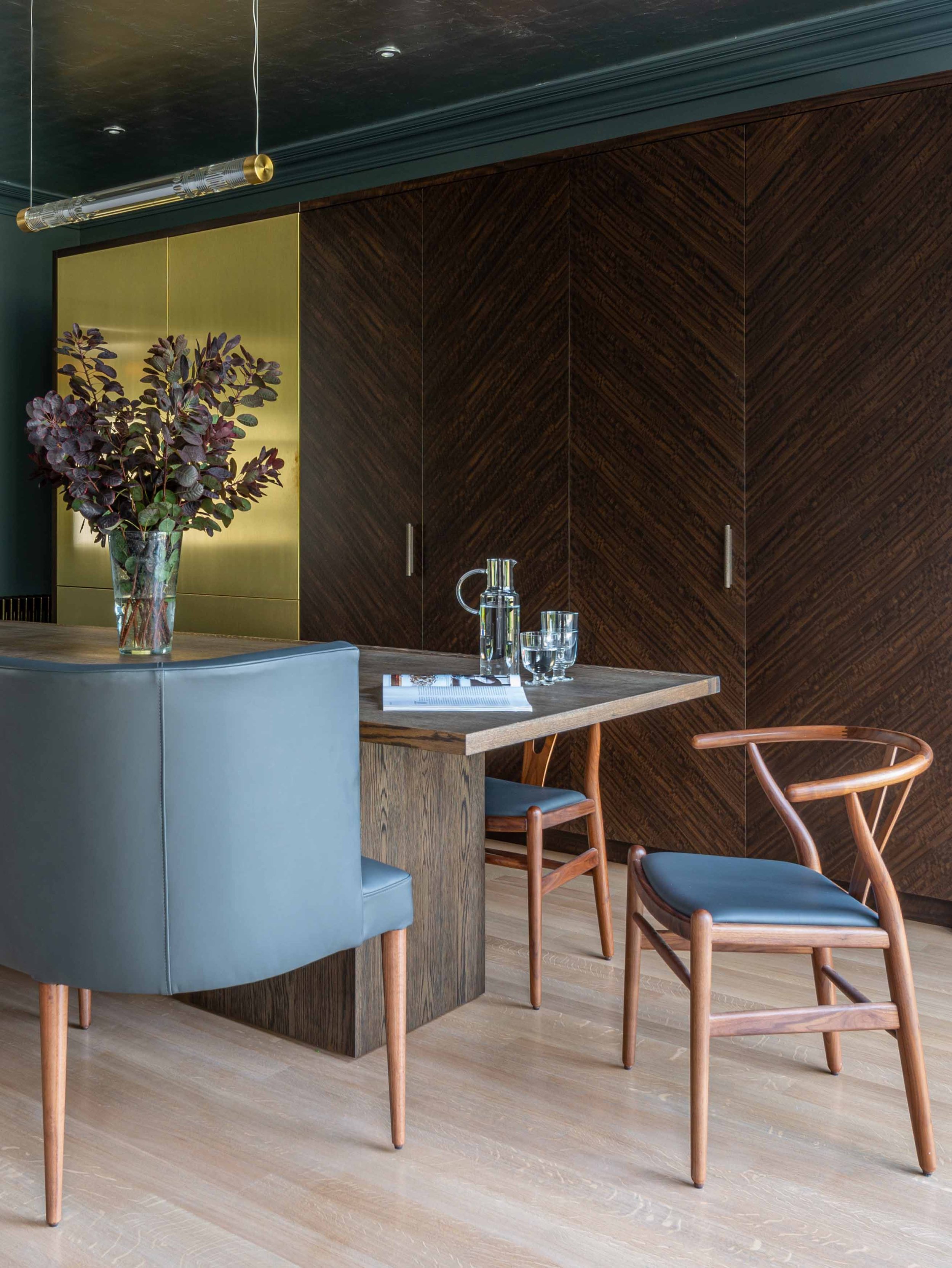 Modern dining room with a wooden table, blue chairs with wooden legs, a glass pitcher with water, drinking glasses, a magazine, and a large vase with dark purple foliage on the table. The room features dark wood paneled cabinets, a gold accent wall, and a green ceiling with recessed lighting and a modern light fixture.