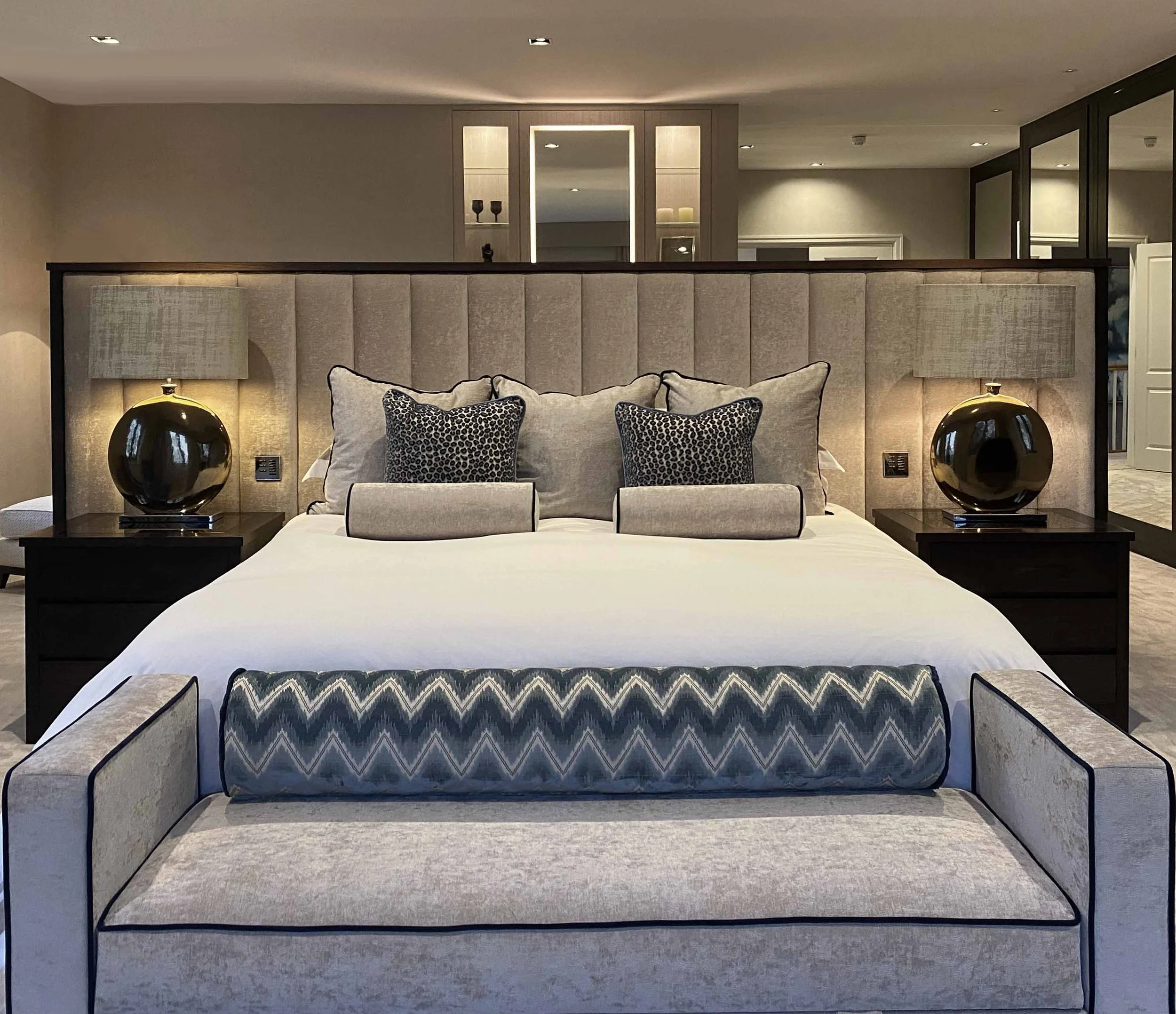 A beautifully designed hotel bedroom with a bed featuring a beige upholstered headboard, surrounded by matching beige and black decor, including bedside lamps, decorative pillows, and a bench at the foot of the bed.