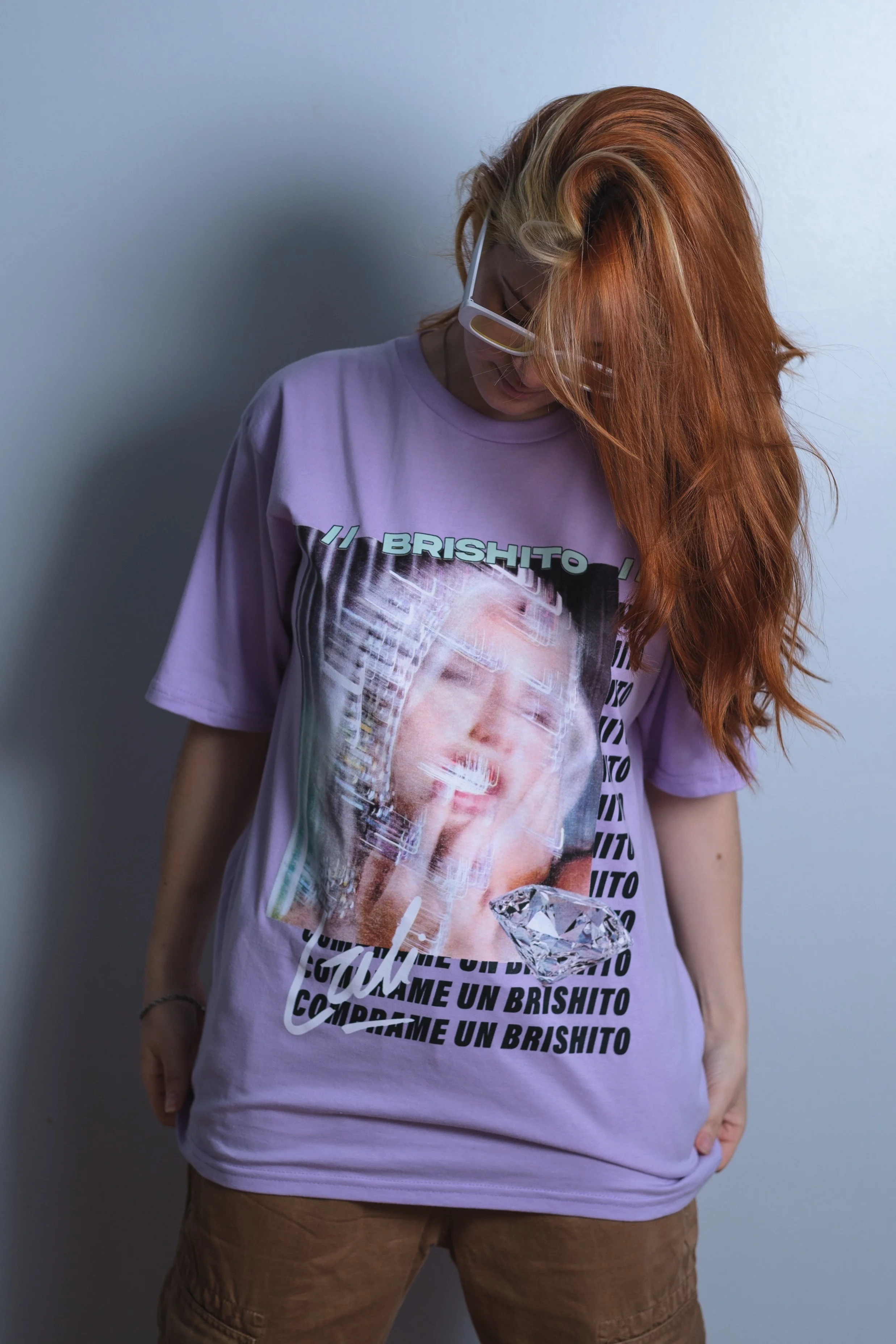 Joven con cabello rojo, gafas blancas, usando una camiseta lila con una imagen de una mujer y palabras en español, frente a una pared blanca.