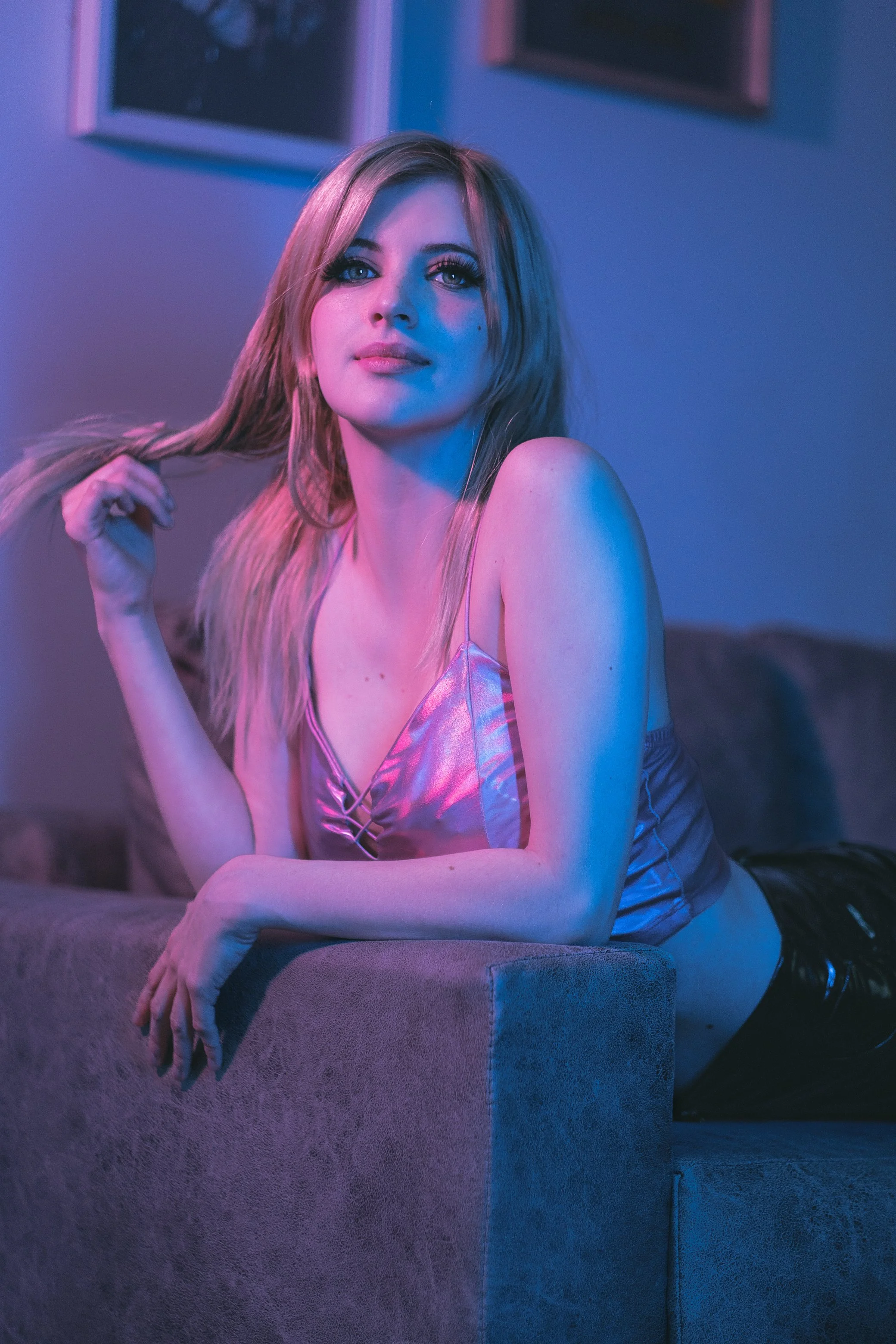 Mujer con cabello rubio, vestido plateado, recostada en un sofá, iluminada con luces de color púrpura y azul en un ambiente moderno.