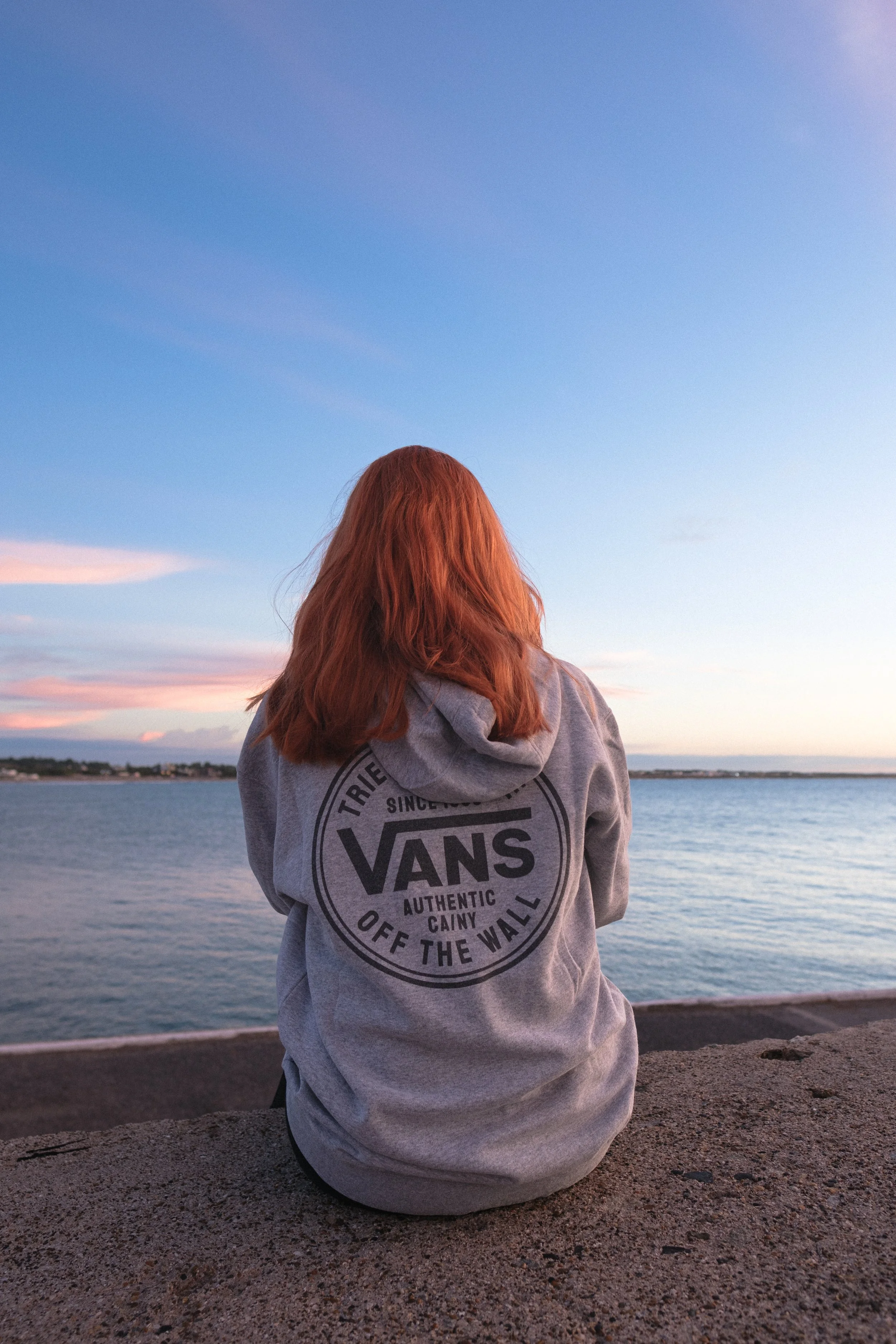 Una persona con cabello rojo sentada junto al mar en una puesta de sol, vistiendo una sudadera gris con logo de Vans en la espalda.