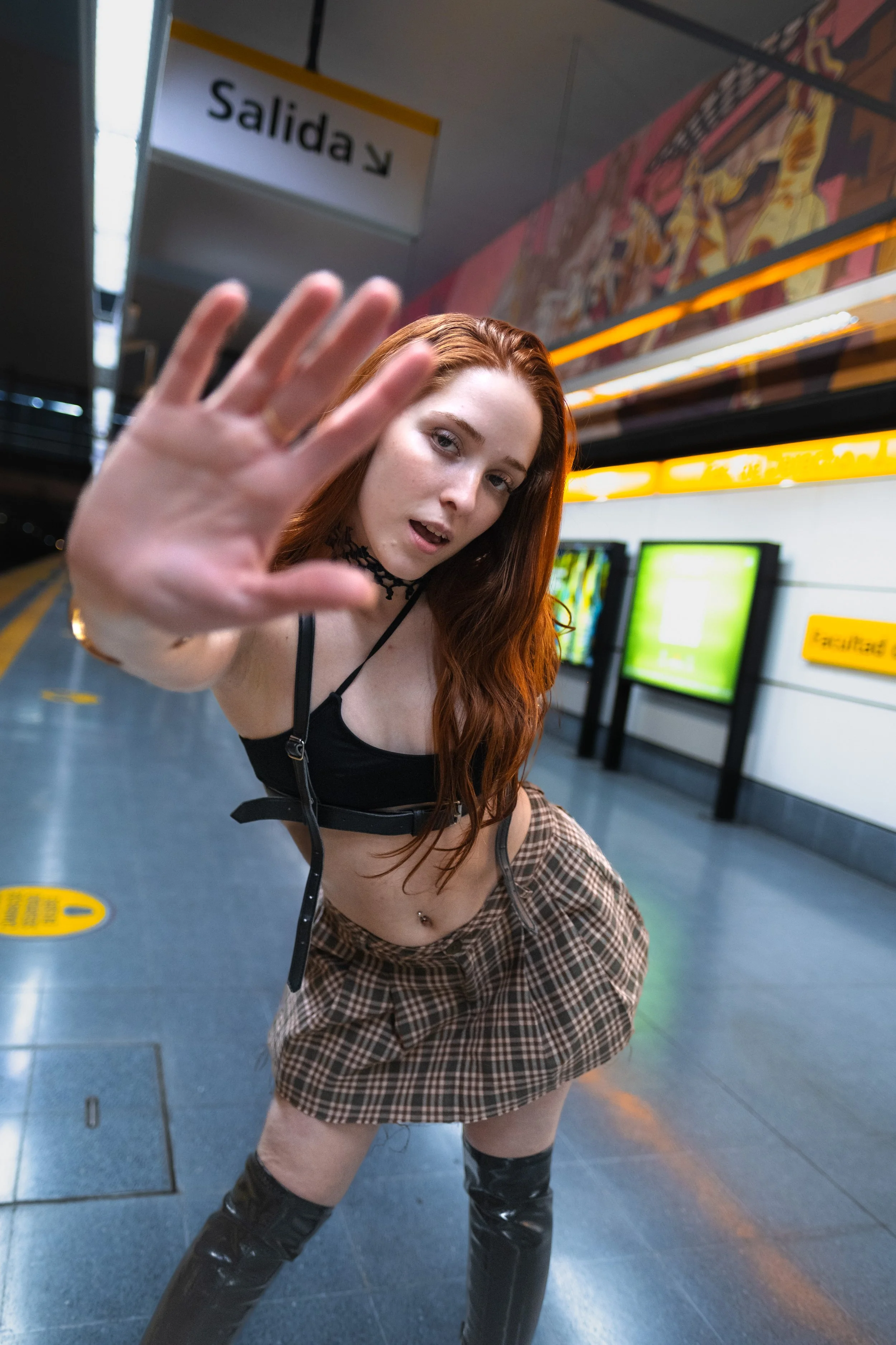 Joven mujer con cabello rizado y piel clara en una estación de metro, con expresión seria y mano extendida hacia la cámara, usando ropa moderna y tacones altos.
