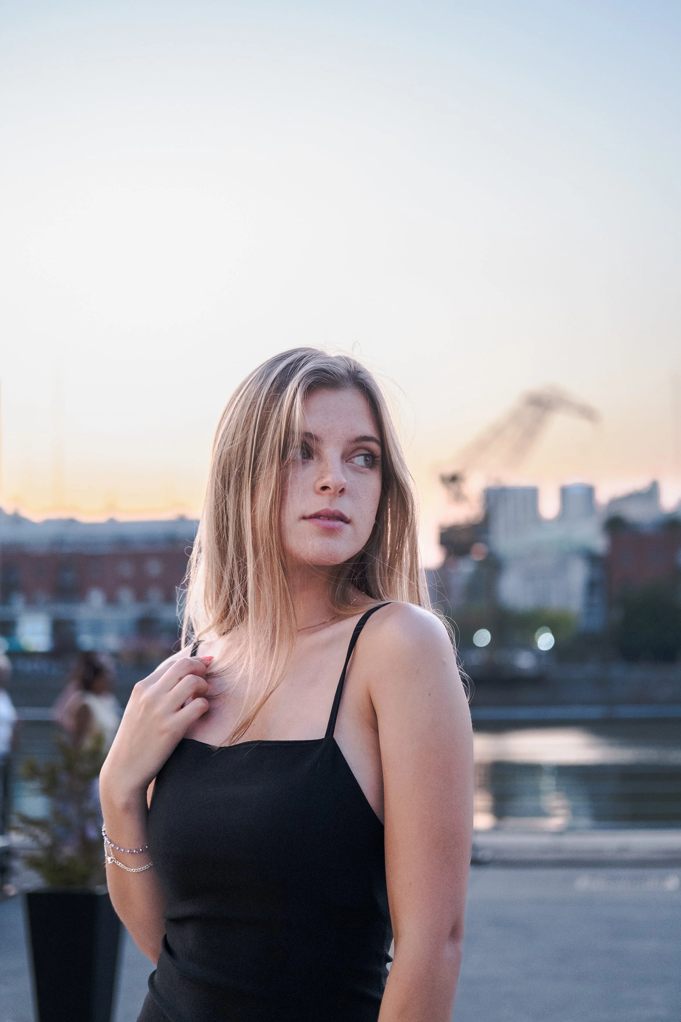 Joven mujer con cabello rubio y suelto, vestido negro de tirantes, en un entorno urbano al atardecer, junto a un río o cuerpo de agua.