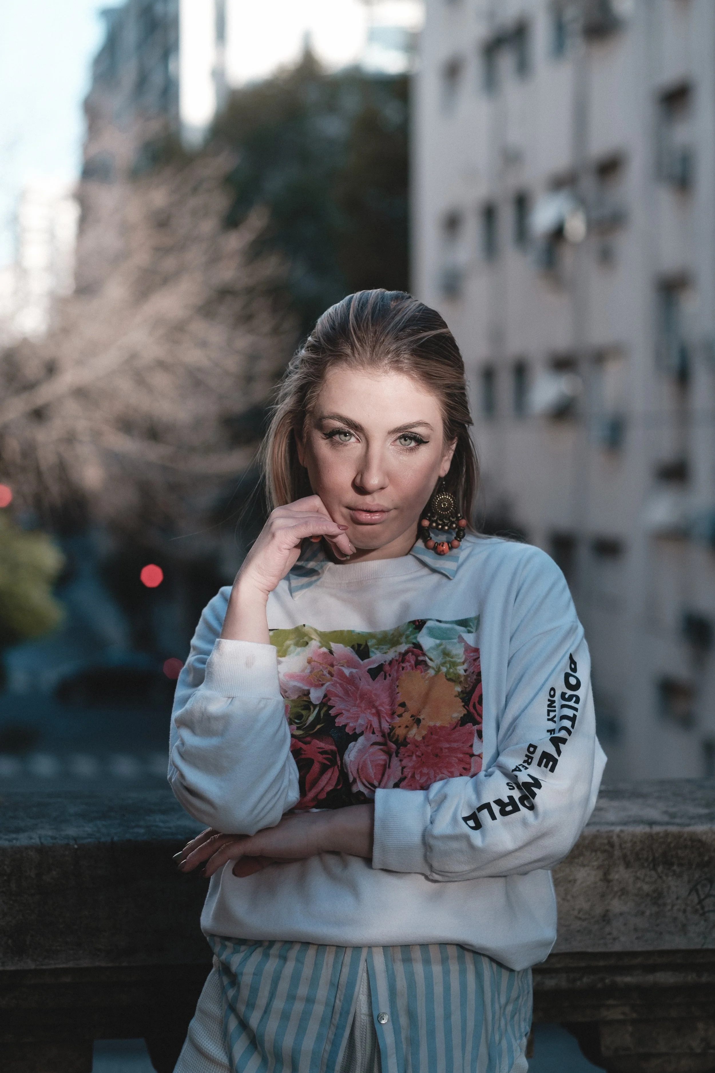 Joven mujer con cabello rubio, usando camiseta blanca con un diseño floral, en un entorno urbano, con edificios y árboles de fondo.