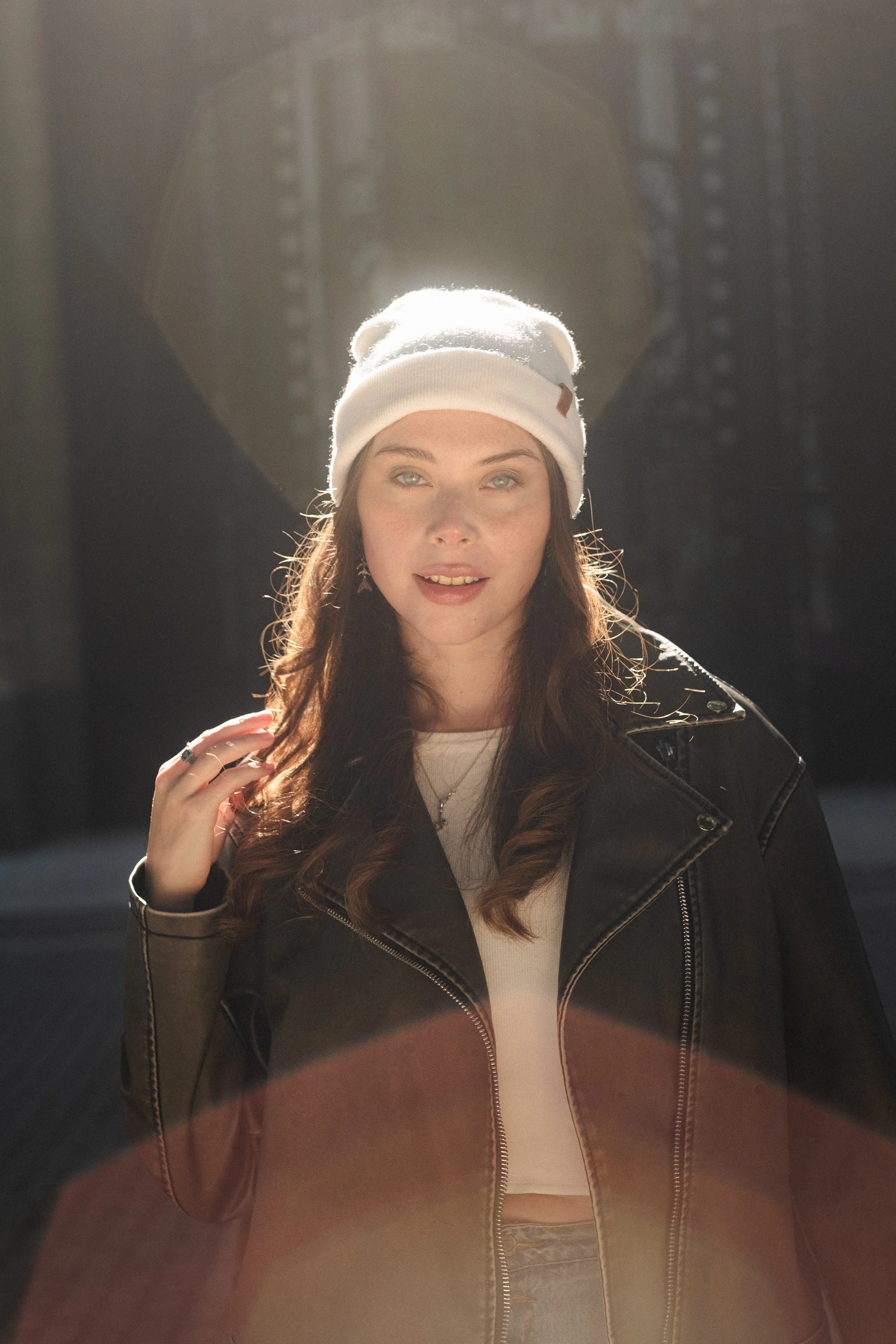 Joven mujer con gorro blanco, chaqueta de cuero y camiseta blanca, posando con luz de sol y fondo urbano.