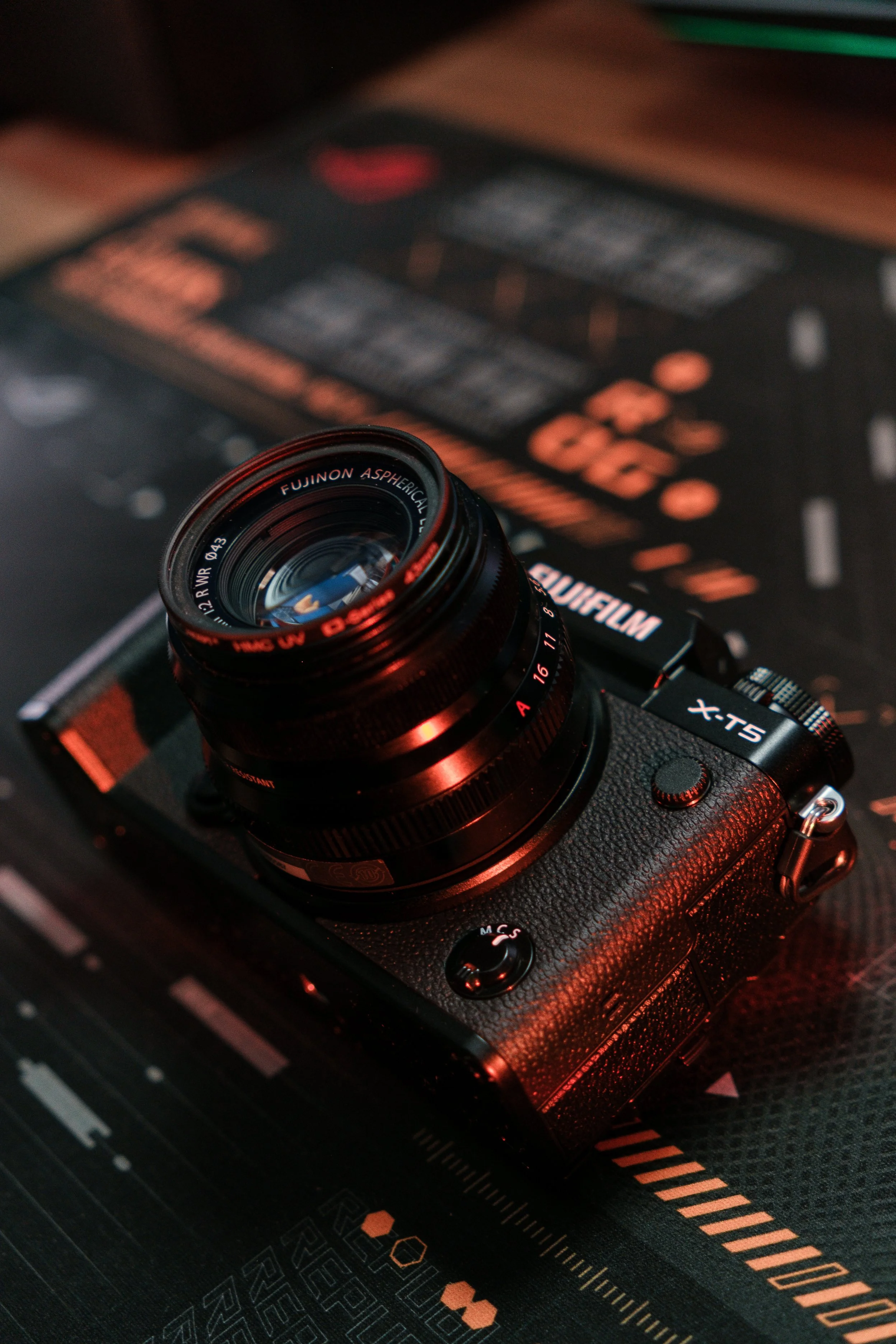 Una cámara fotográfica Fujifilm X-T5 con lente Fujinon en primer plano, sobre una superficie negra con detalles en naranja y esquinas curvas.