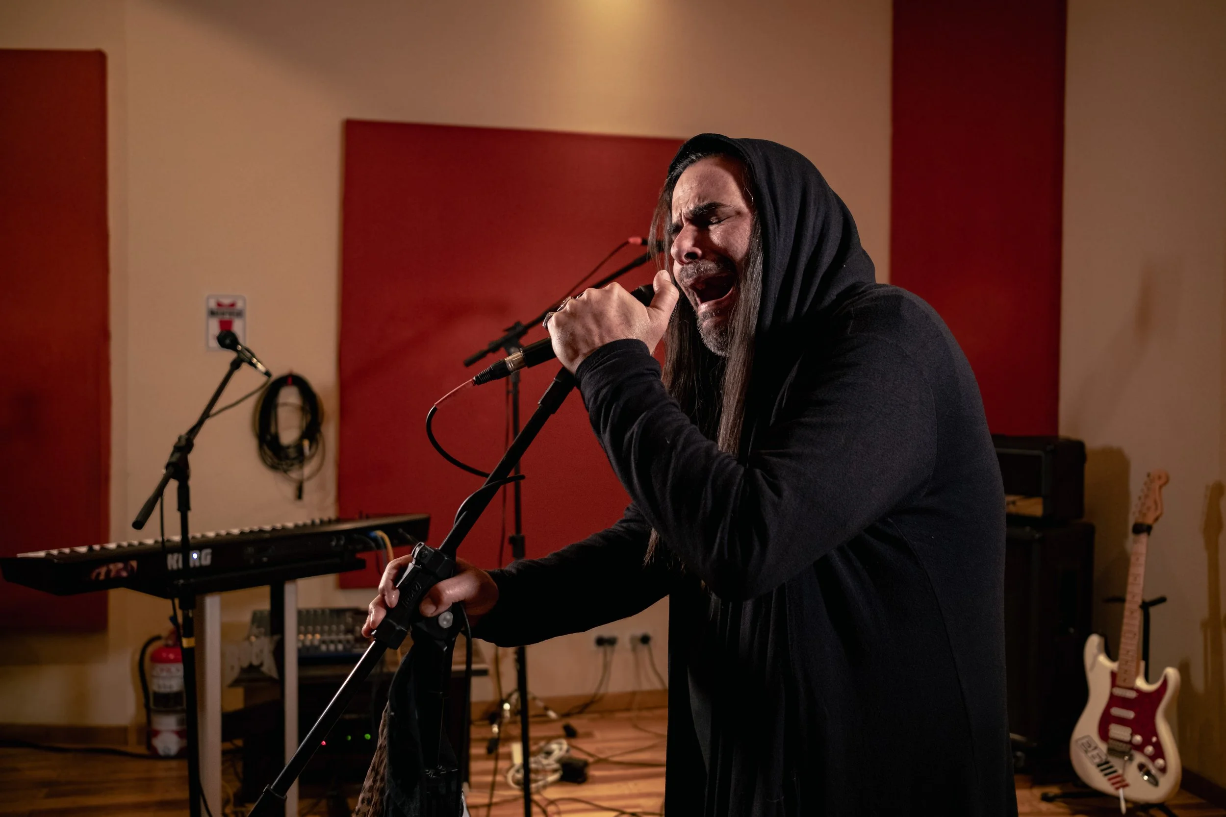 Hombre cantando en un estudio de música, con equipo musical y guitarra eléctrica en el fondo.