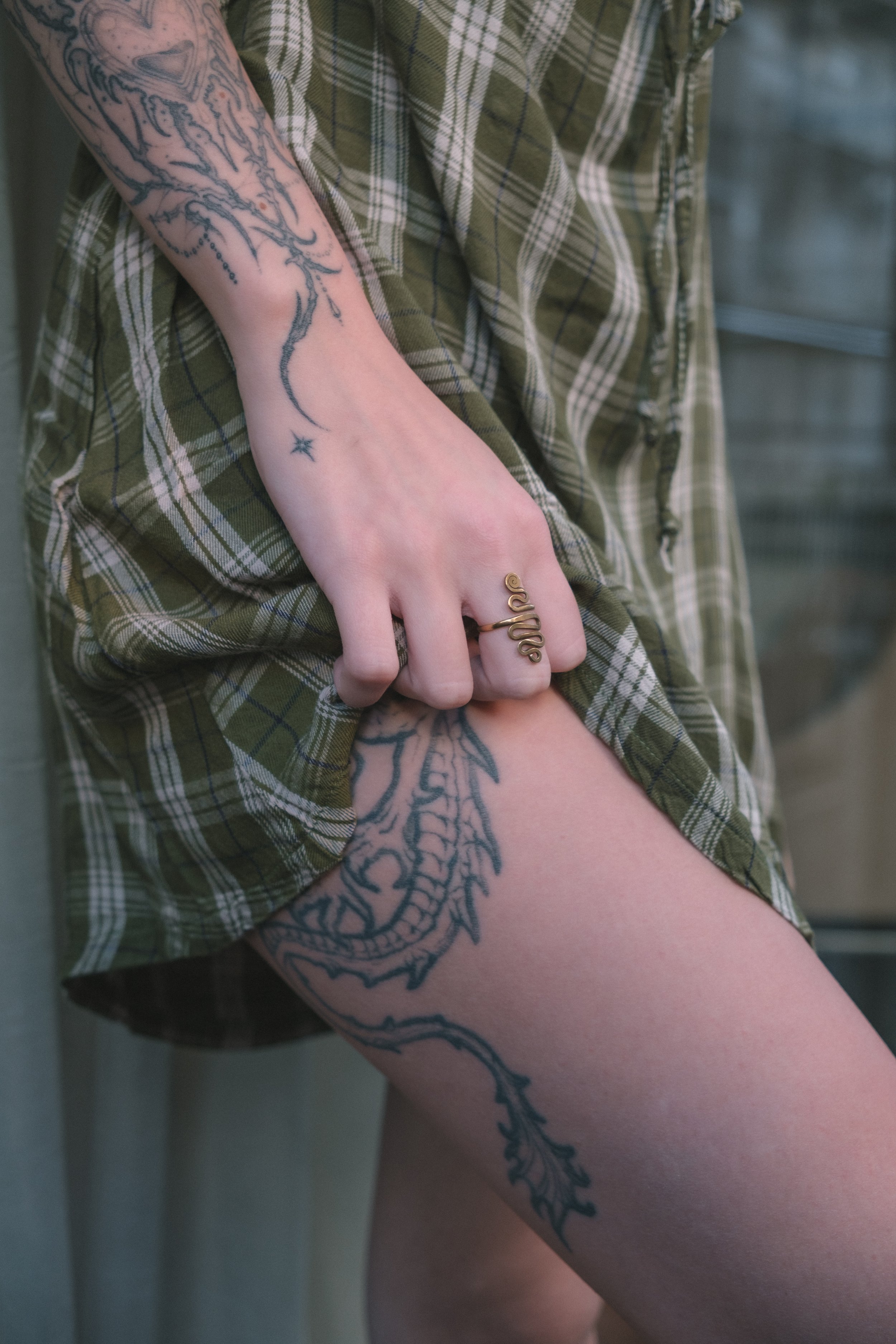 Persona con tatuajes en brazo y pierna, sosteniendo una prenda de ropa de cuadros de color verde y beige.
