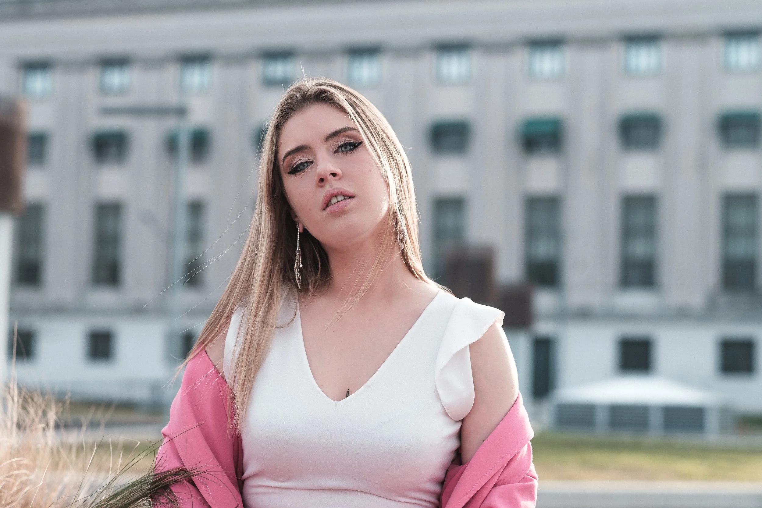 Joven mujer con cabello rubio, vistiendo blusa blanca y saco rosa, en un fondo de edificio moderno.