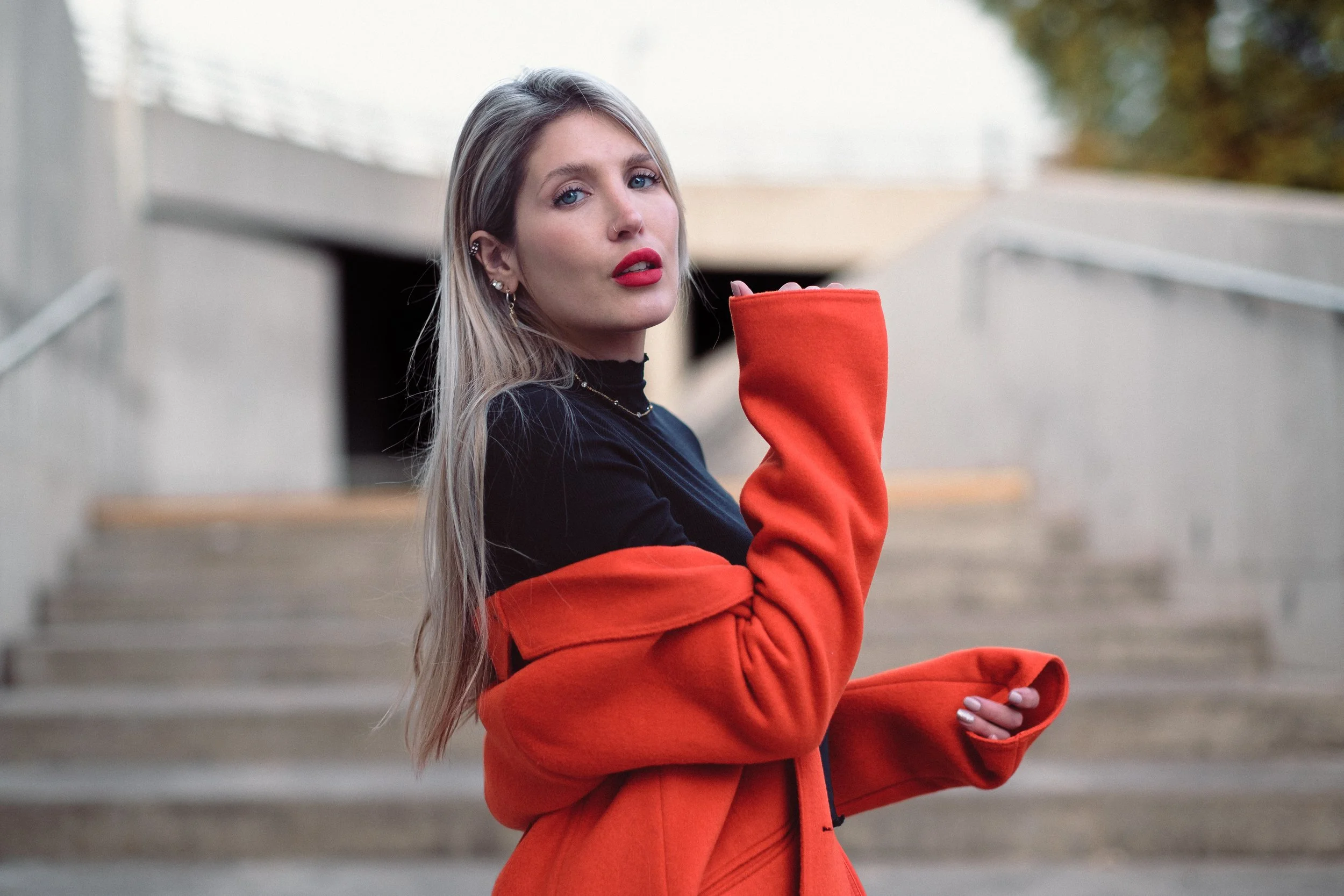 Joven mujer con cabello rubio largo, labios rojos y accesorios de oro en un entorno urbano con escaleras de concreto.