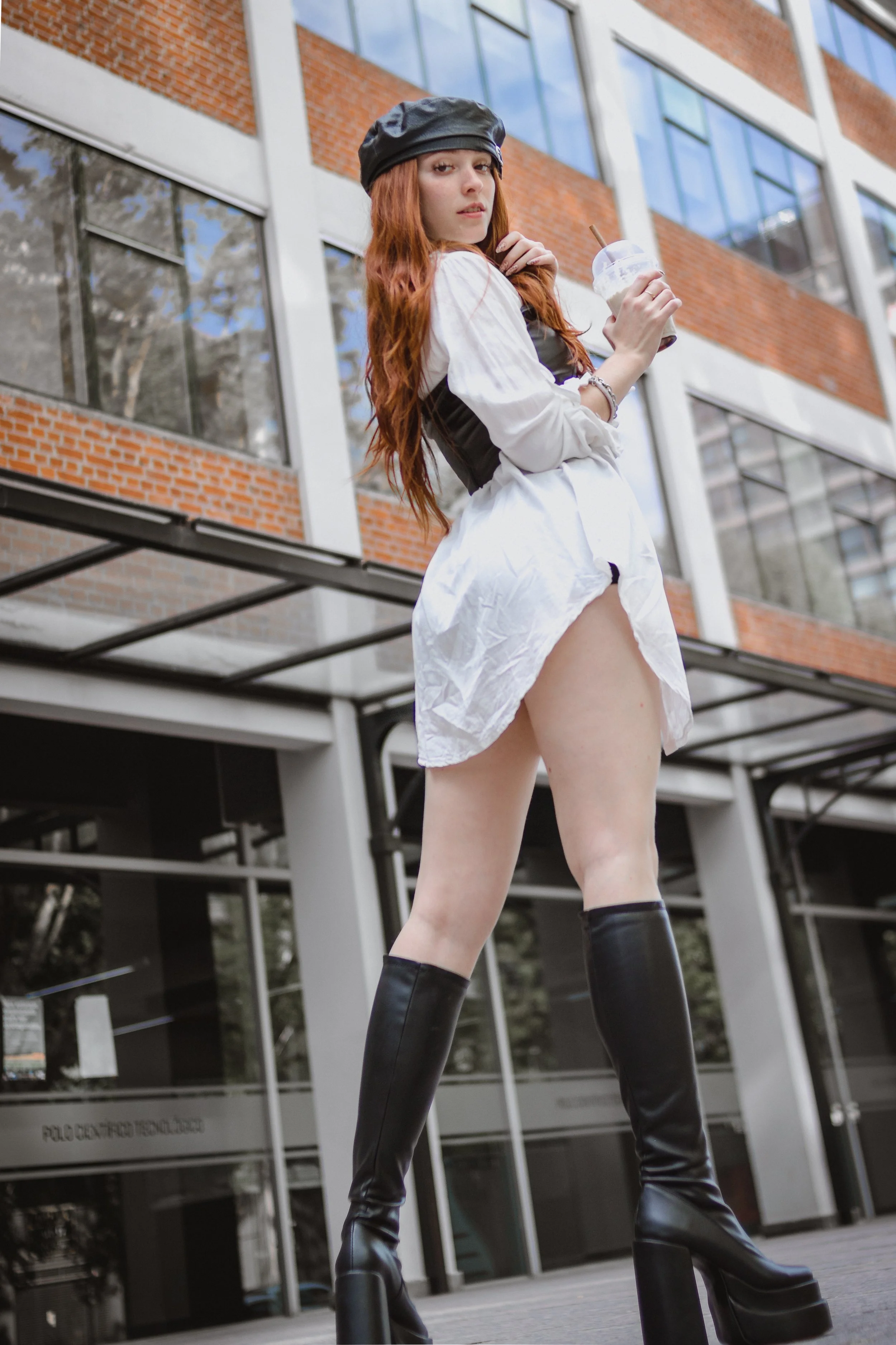Mujer con botas altas negras, vestido blanco, blusa blanca con corsé negro, sombrero negro, sosteniendo un helado, frente a un edificio moderno con ventanas de vidrio y paredes de ladrillo.