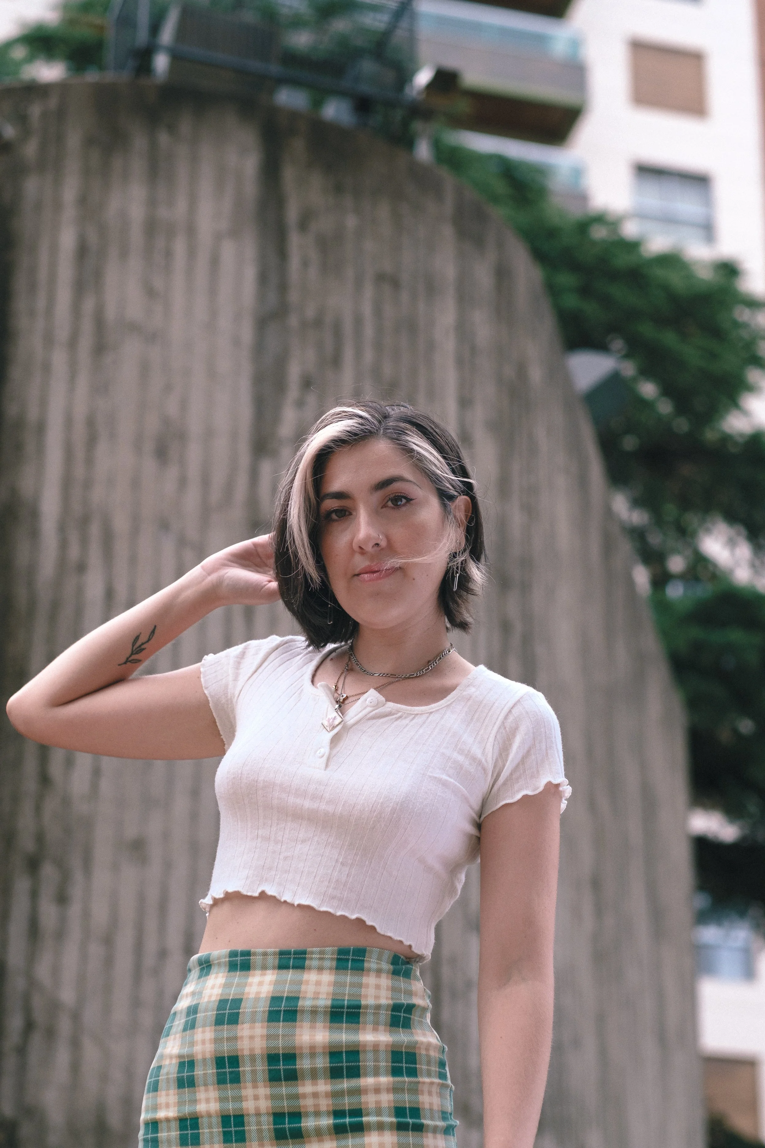 Joven mujer con cabello corto y maquillaje natural, posando frente a un muro de concreto en una ciudad, viste una camiseta blanca ajustada y una falda de cuadros.