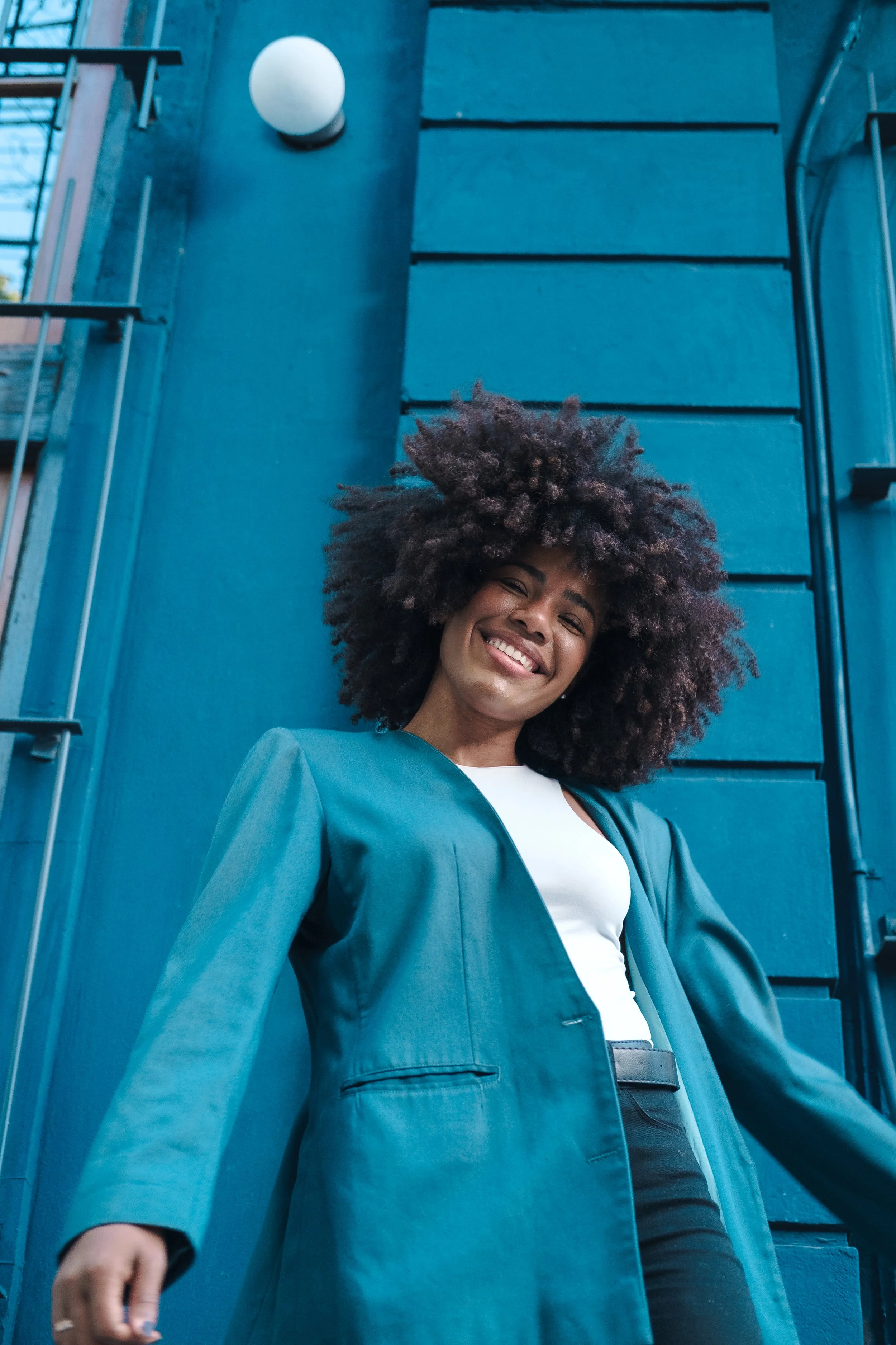 Mujer sonriente con cabello rizado oscuro, vestida con saco azul y camiseta blanca, de pie frente a una pared azul.