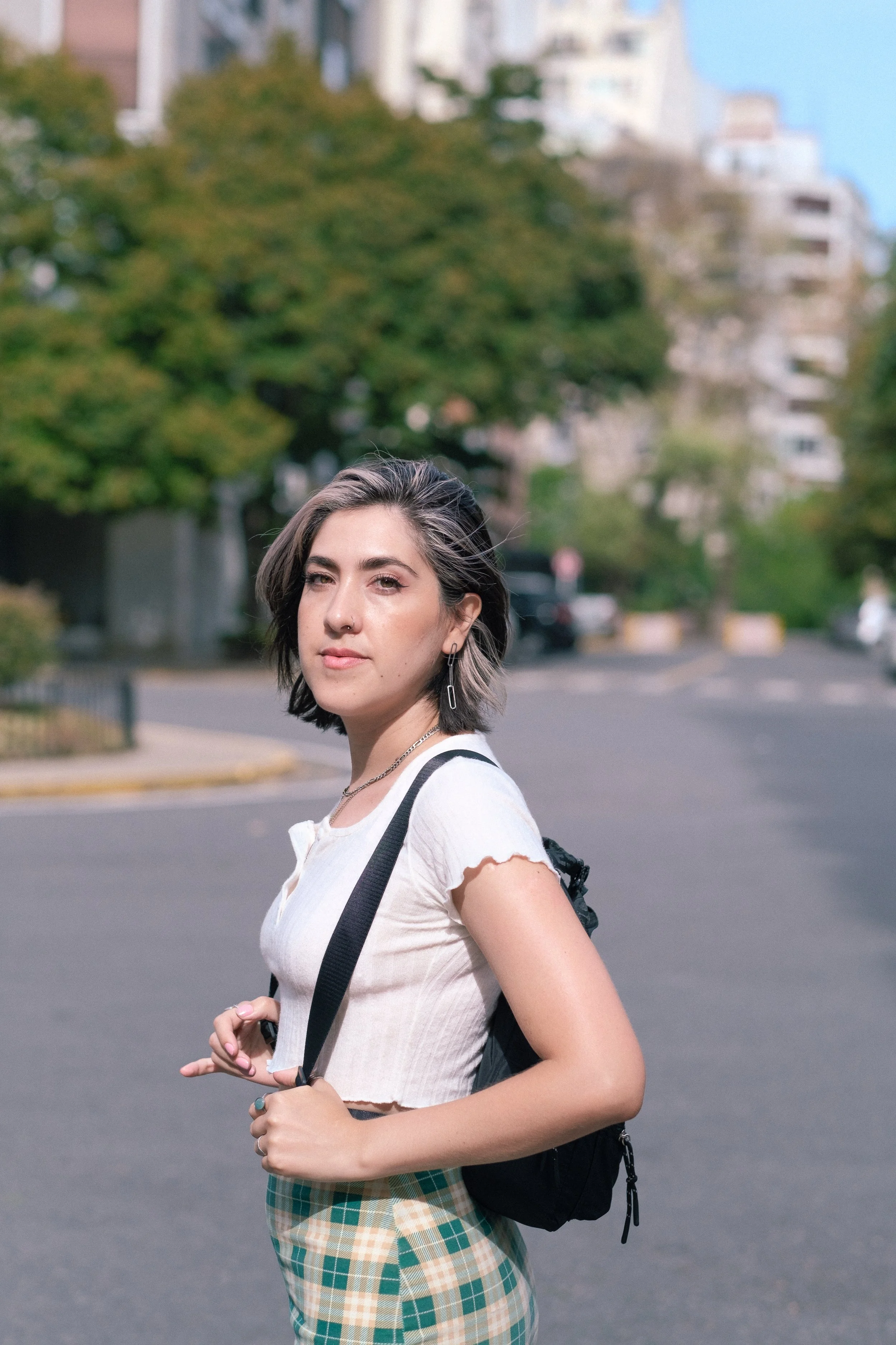 Joven mujer con cabello castaño y arreglado, vestida con blusa blanca y falda a cuadros, lleva mochila negra y mira hacia la cámara en una calle con árboles y edificios al fondo.