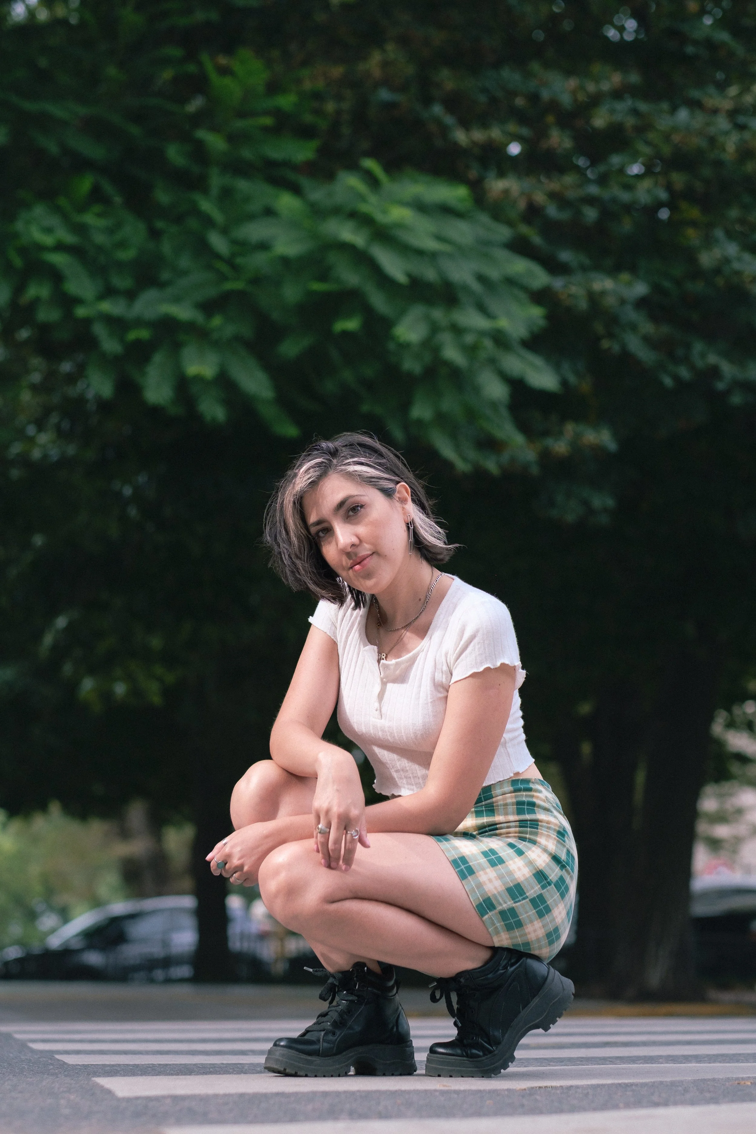 Una joven en pose agachada con vestimenta casual en la calle, con árboles verdes en el fondo.