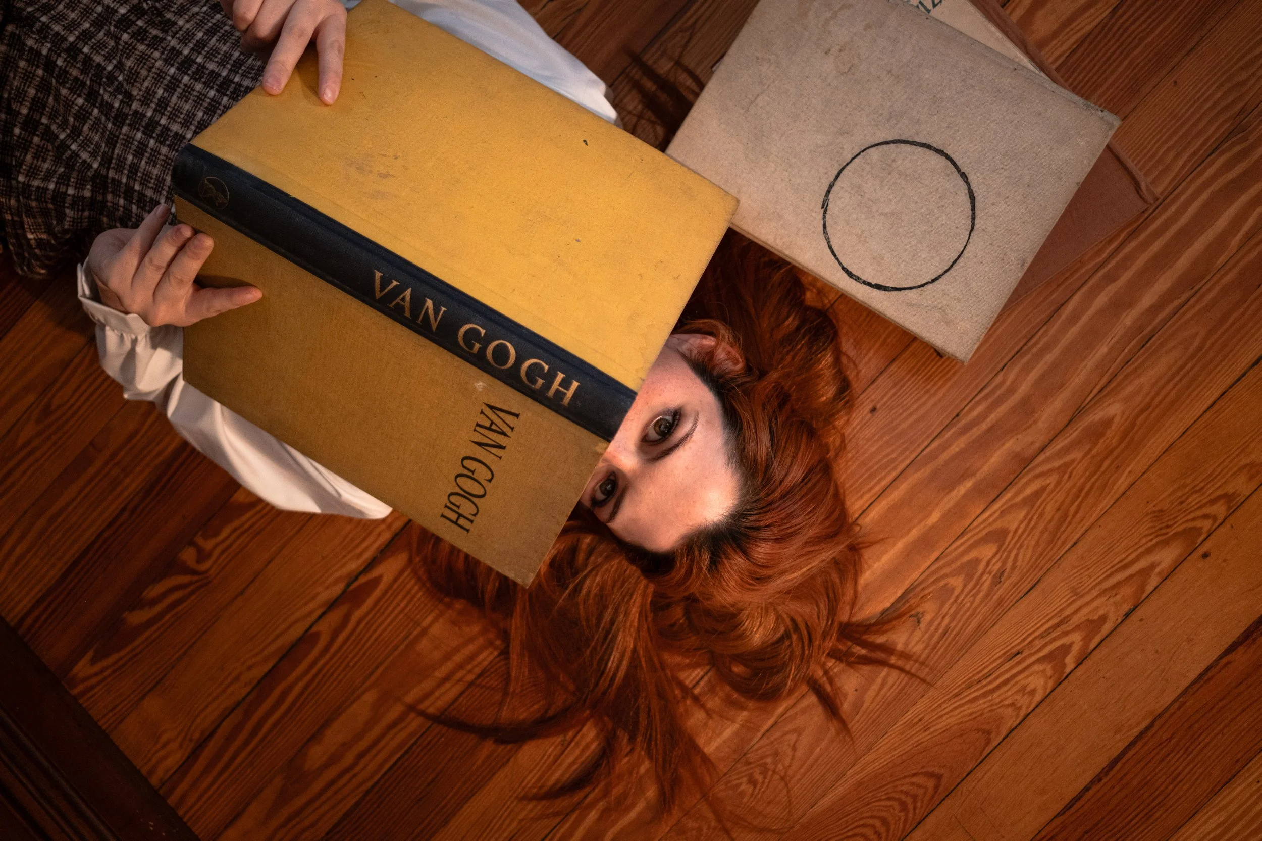 Una mujer con cabello rizado y pelirrojo, acostada en un suelo de madera, sosteniendo un libro grande de tapa dura con el título 'VAN GOGH'. La mujer mira hacia arriba, con su rostro parcialmente oculto por el libro. A su lado hay un cuadro en blanco con un círculo dibujado en el centro.