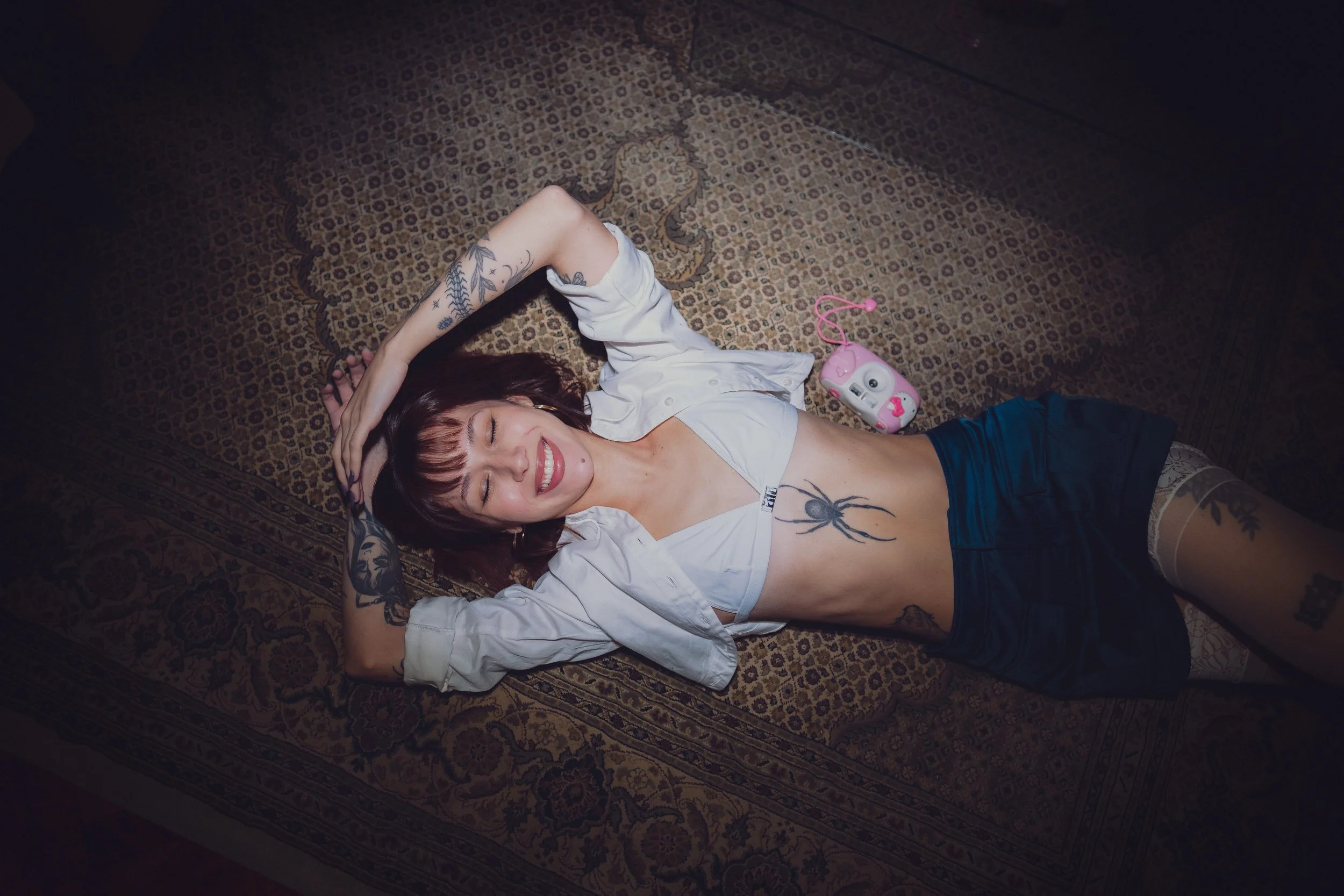 Mujer joven con tatuajes, acostada en una alfombra, sonriendo, con una camiseta blanca, pantalones cortos negros, y un juguete rosa junto a ella.
