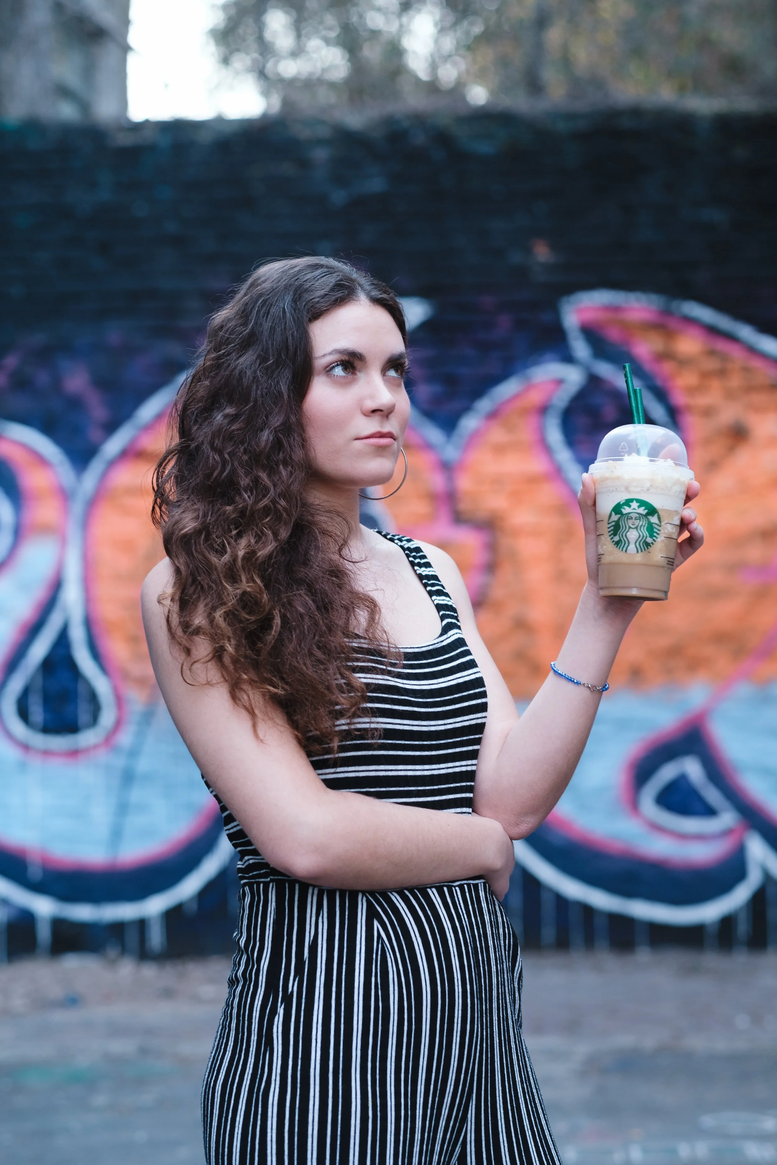 Una joven con cabello rizado, vestida con un atuendo a rayas, sostiene una bebida fría de Starbucks en un entorno urbano con un mural de graffiti en el fondo.