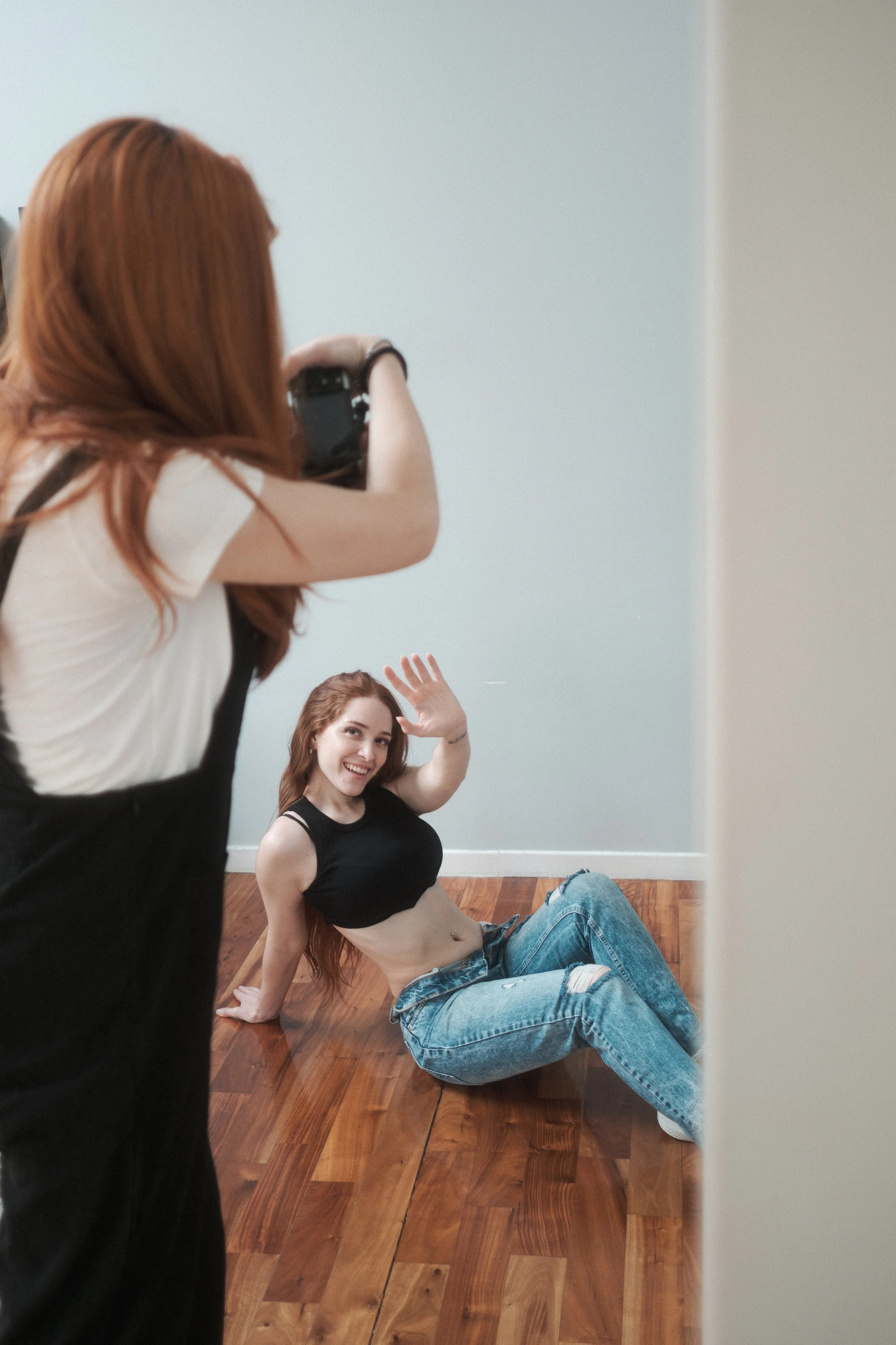 Una joven de cabello largo y castaño claro posa en una sesión de fotografía, sentada en el suelo de madera con una camiseta negra y jeans rotos, sonriendo y saludando a la cámara.