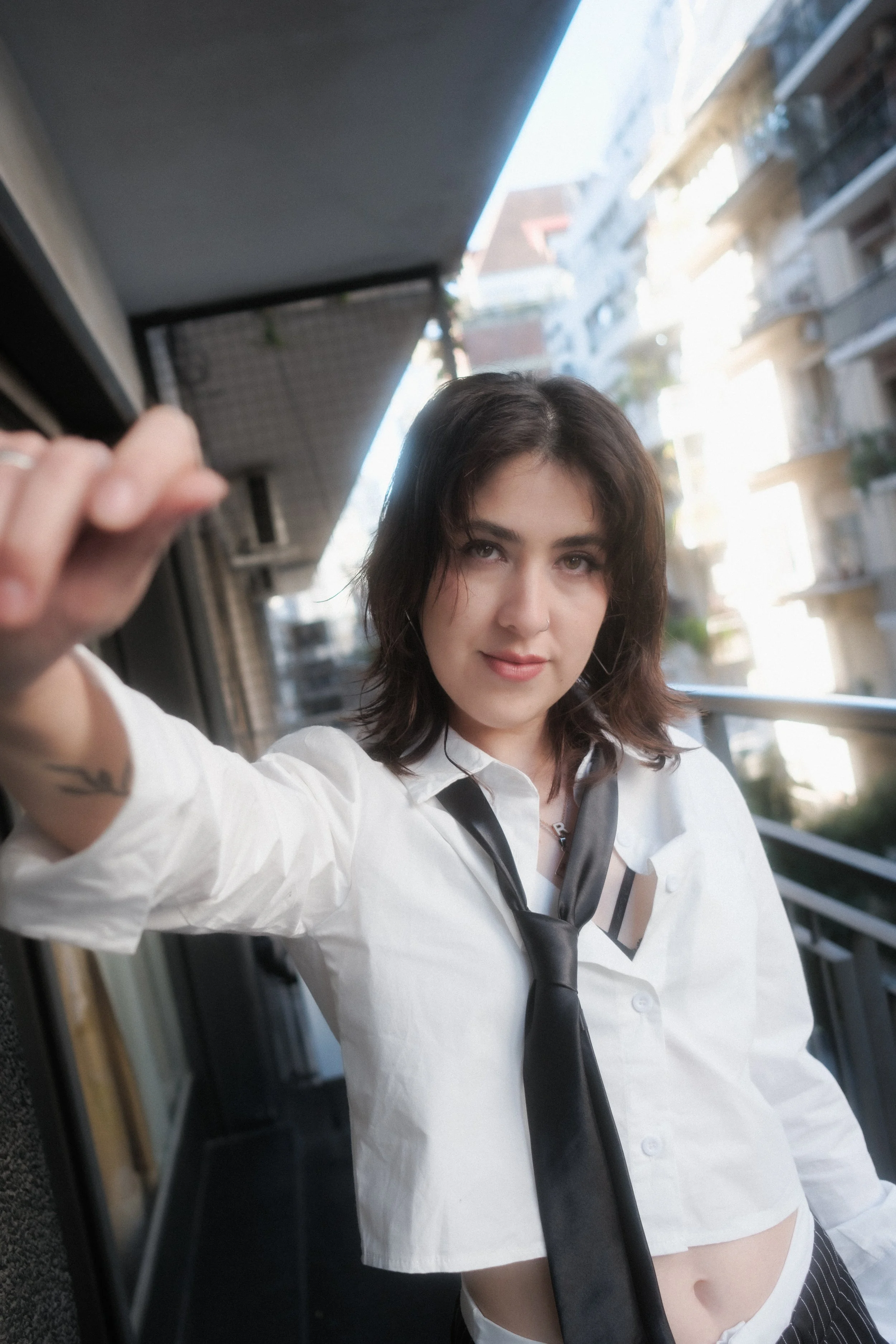 Juvenil mujer con cabello rubio oscuro, blusa blanca con manga larga y corbata negra, posando en un balcón en una ciudad de edificios altos, con expresión confiada.