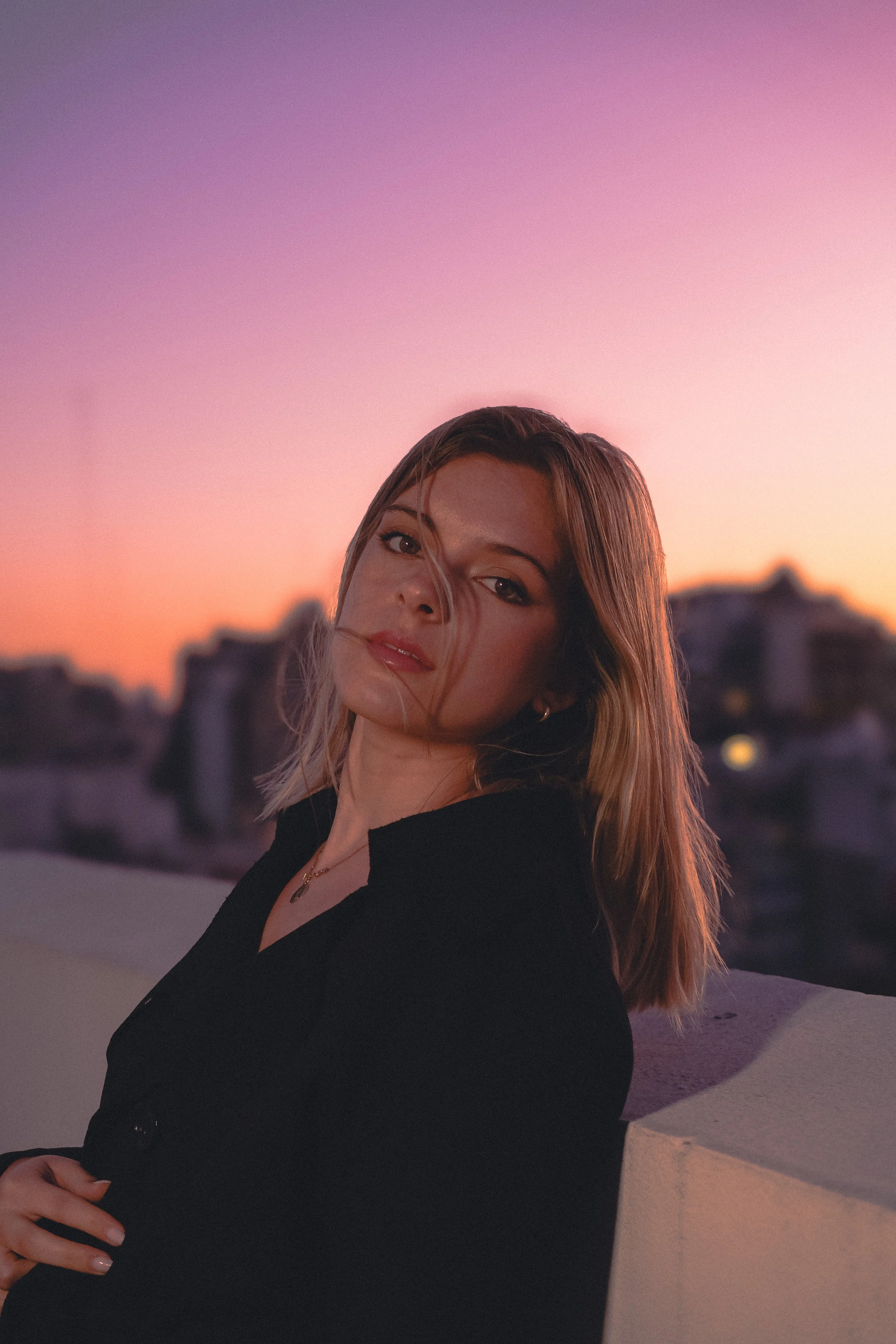 Mujer con cabello rubio y suelto, usa una blusa negra y está recargada en una pared, en un atardecer con cielo colorido en el fondo.