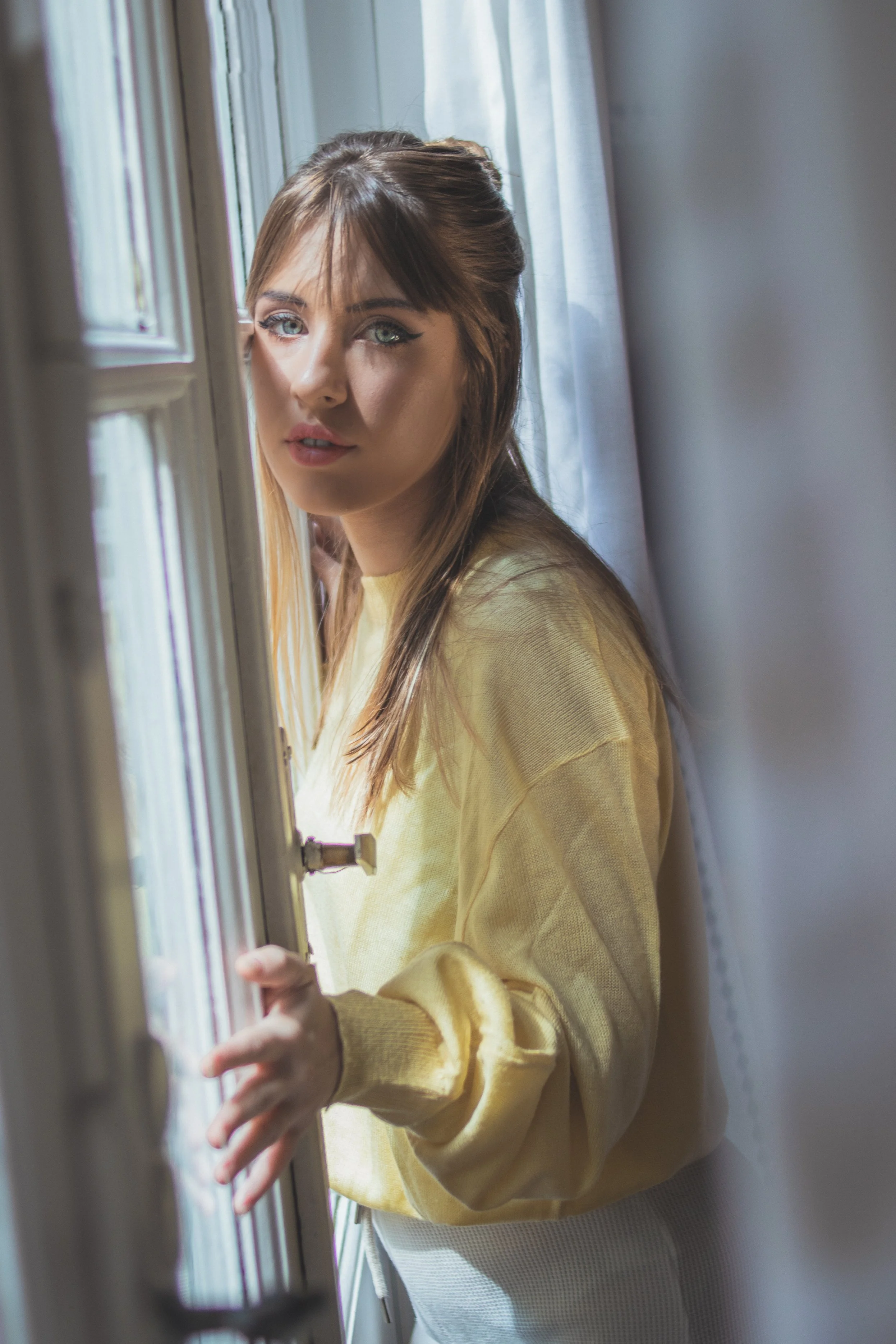 Joven mujer con cabello castaño y ojos azules, usando un suéter amarillo, mirando por una ventana en una habitación con cortinas blancas.
