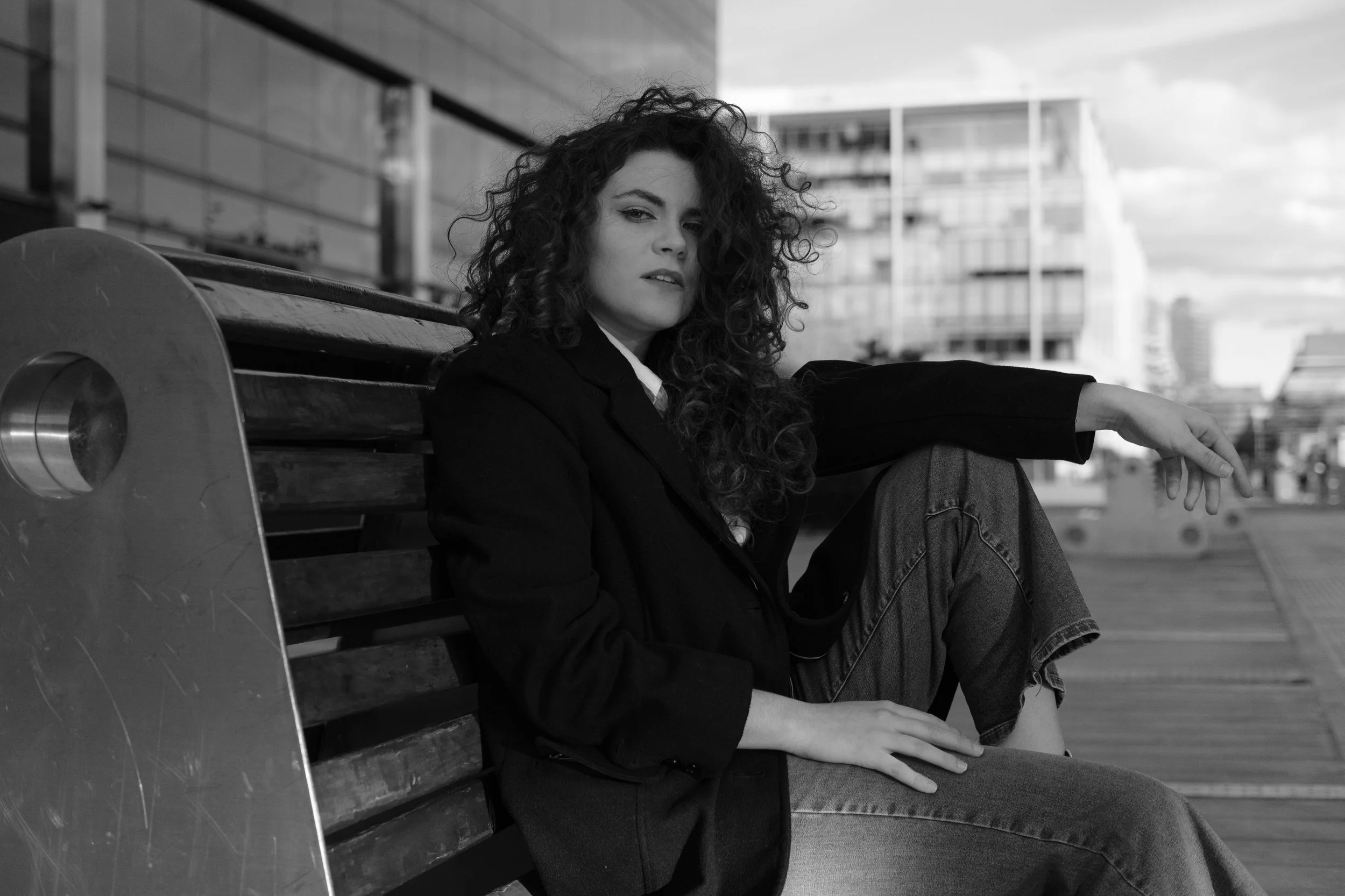 Mujer con cabello rizado sentado en un banco en un entorno urbano en blanco y negro.