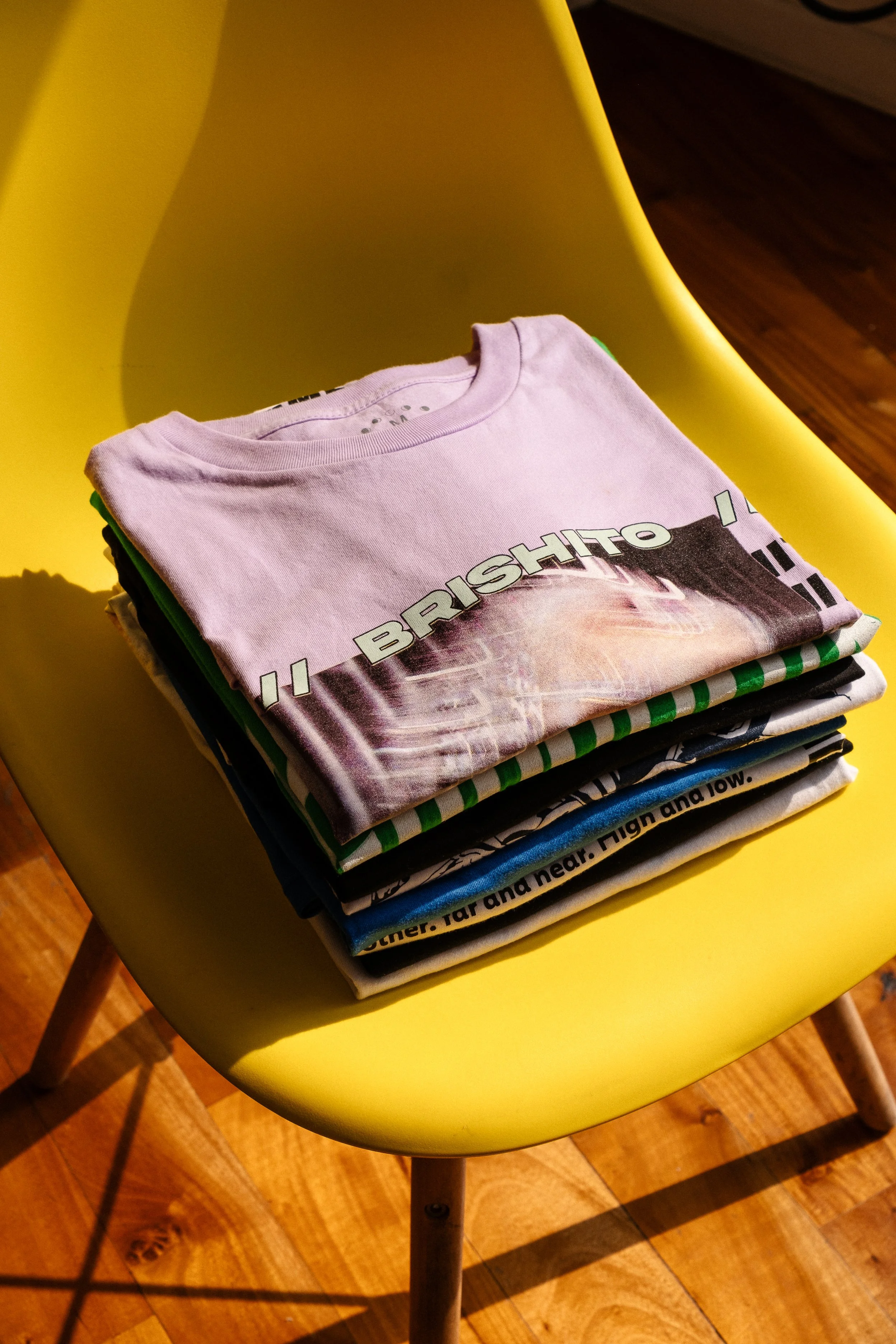 Una silla amarilla con una pila de camisetas apiladas sobre ella, incluyendo una con el texto "BRISHITO" y otras con diferentes diseños.