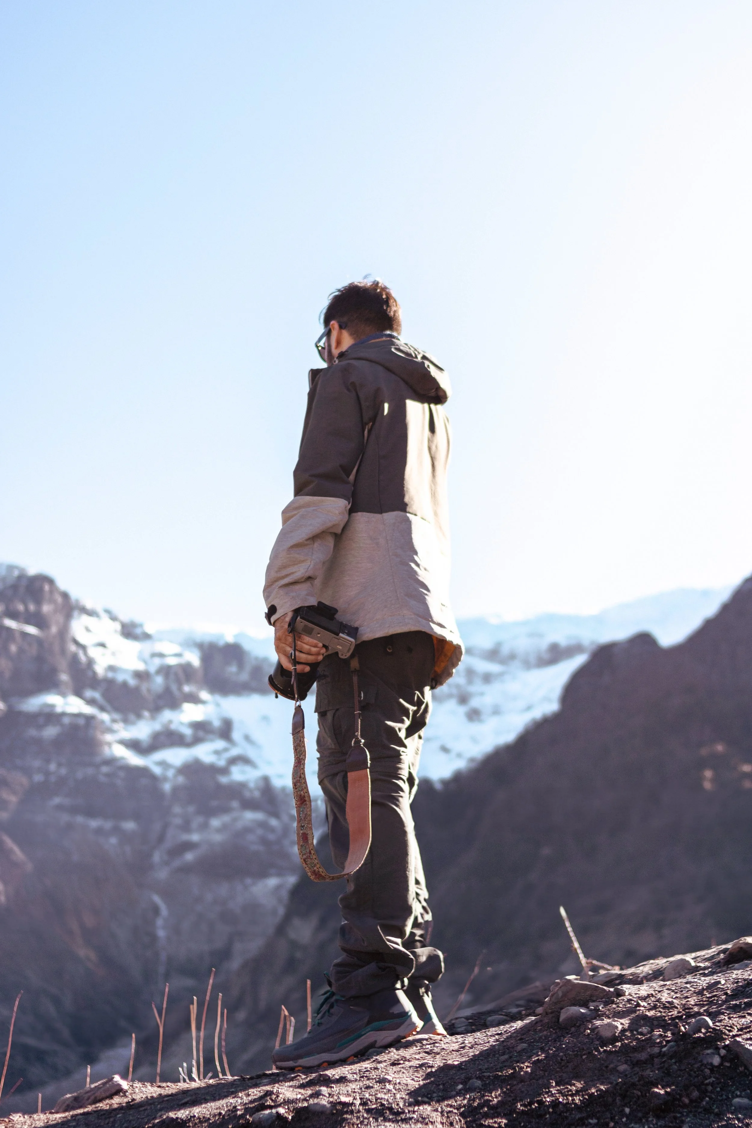 Persona con chaqueta y pantalones de senderismo sostiene una cámara en un paisaje montañoso con nieve y rocas.